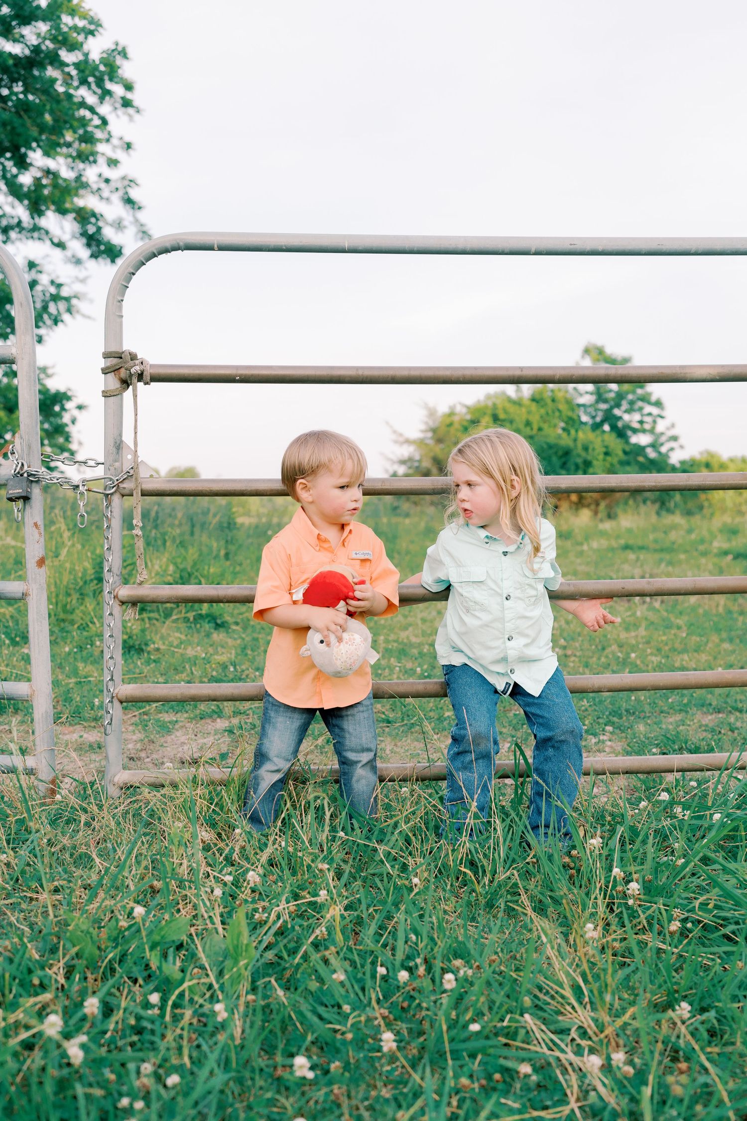 Two children stand by metal fence gate holding toys in a rural setting.