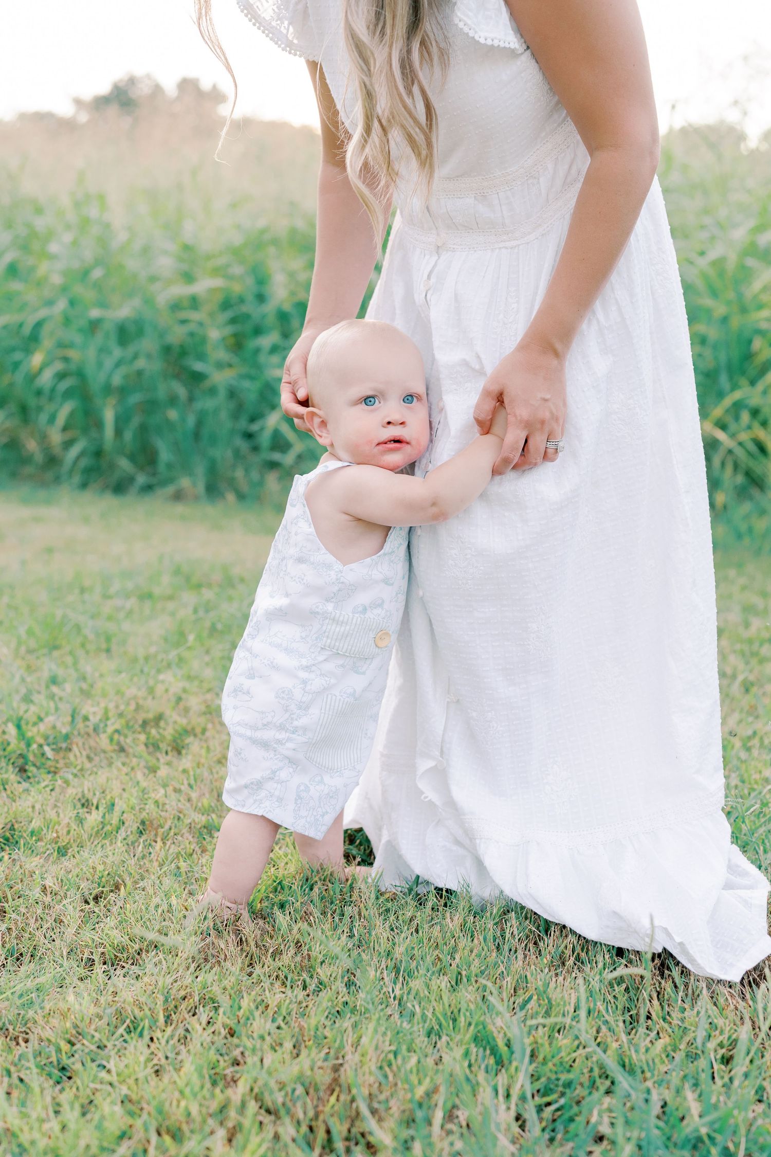 Close-up shots of a baby in white clothing taking first steps with support in a summer field.