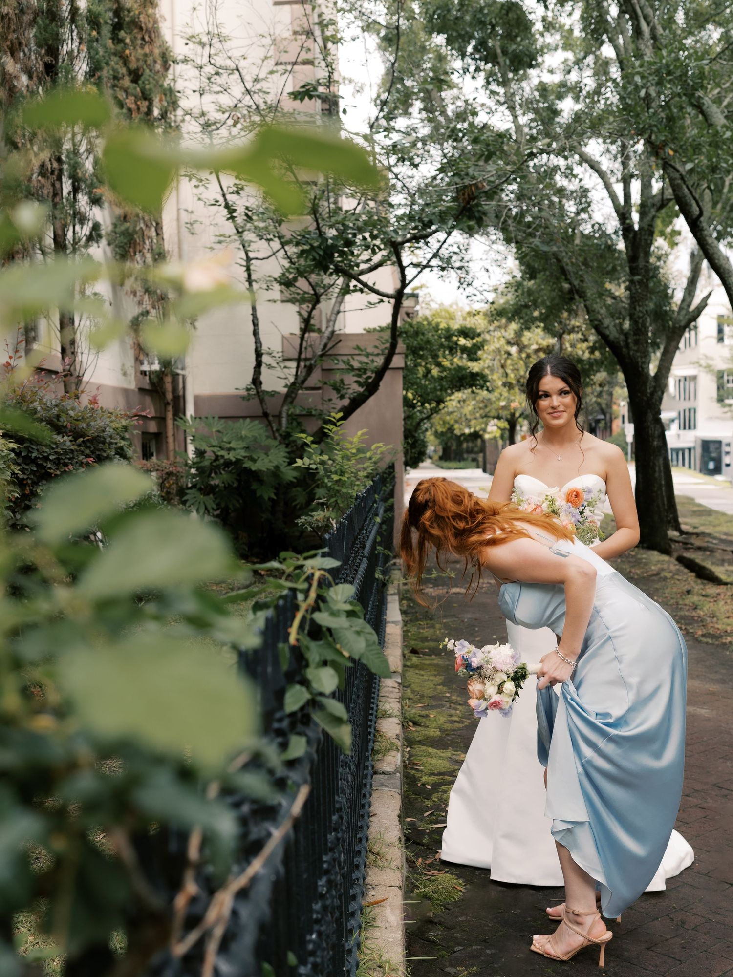 A wedding party in light blue dresses poses together in a garden setting with trees and greenery.