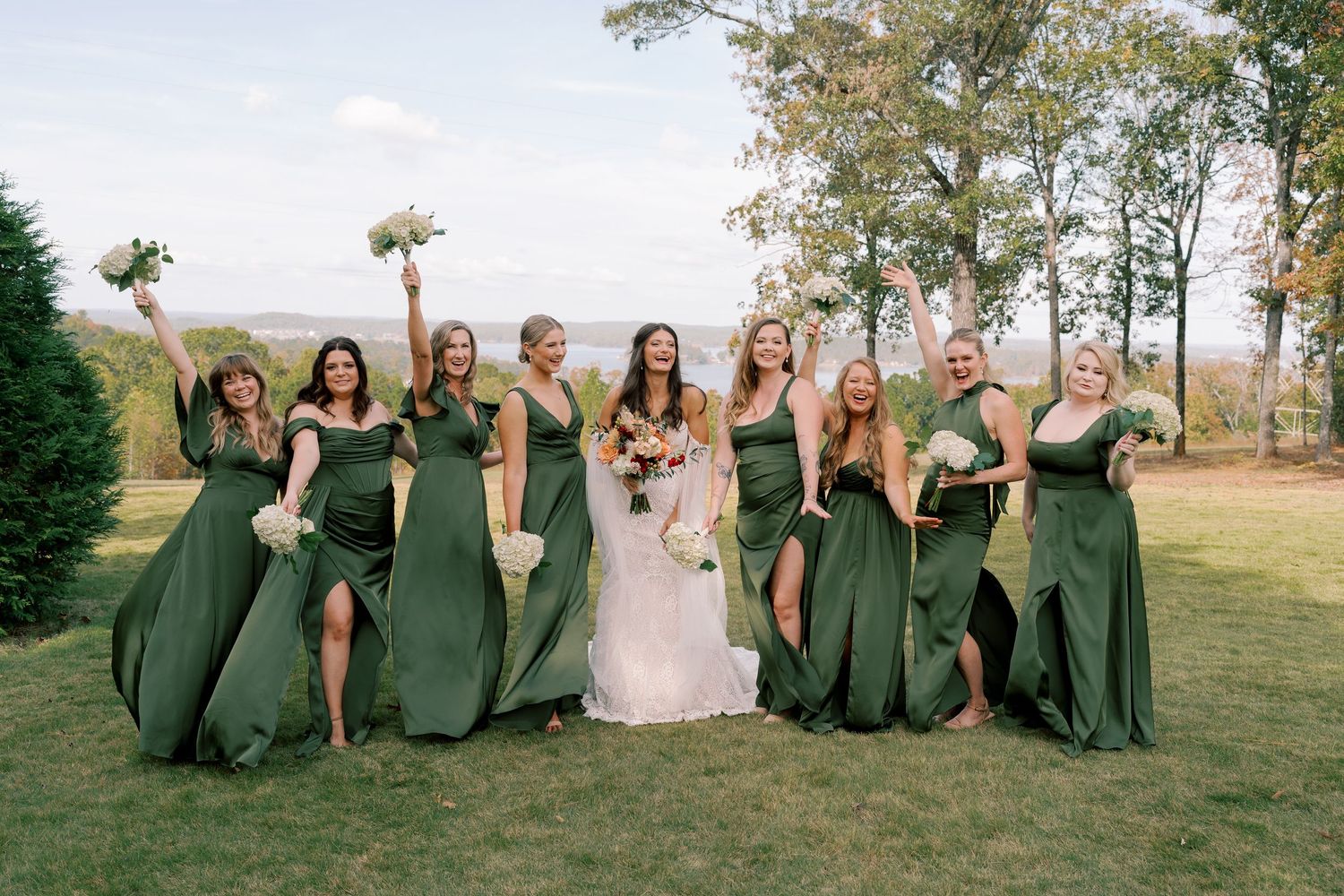 Wedding party in emerald green dresses poses playfully on a grassy hillside.