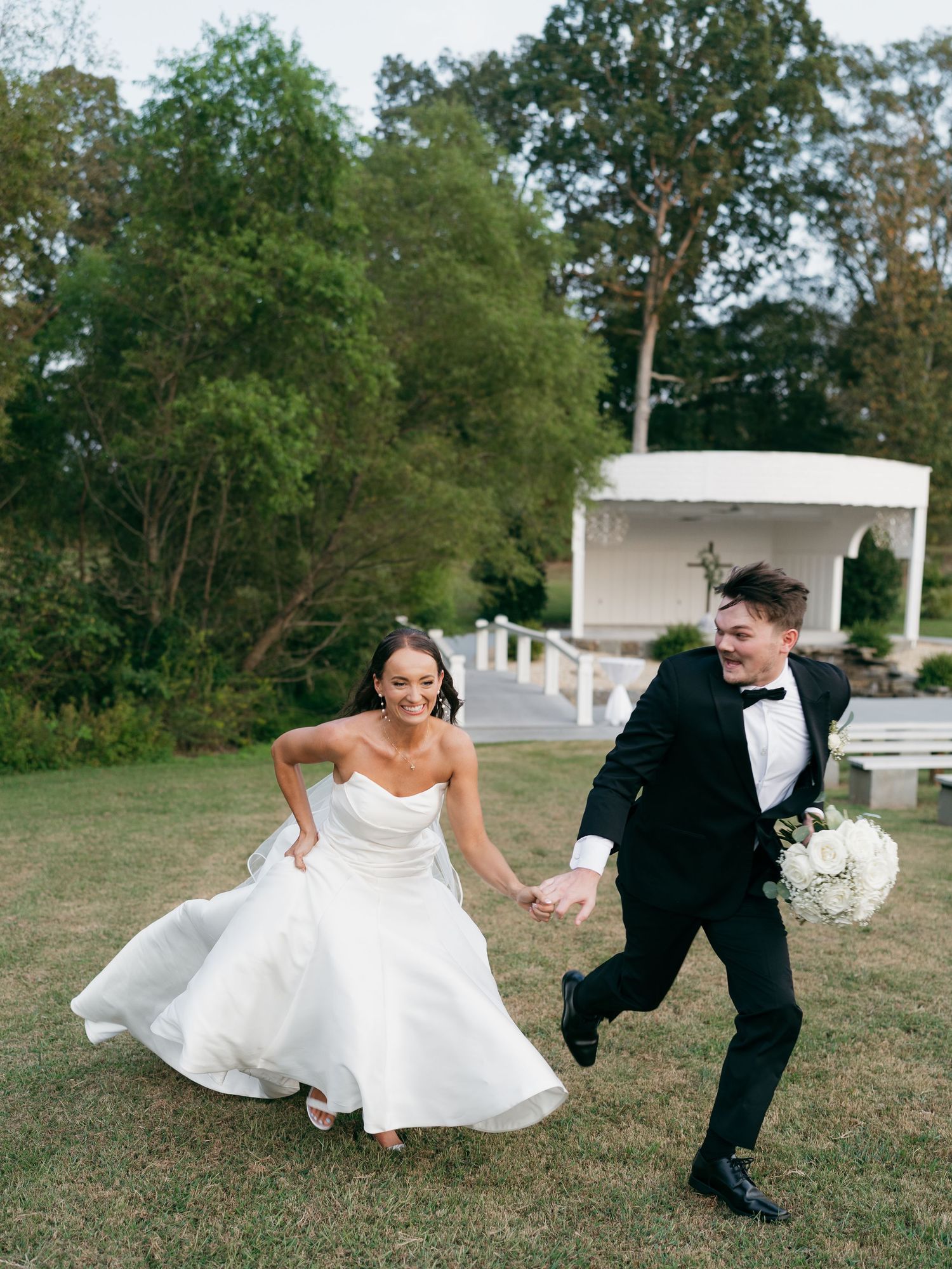 Playful wedding couple running and laughing together on grassy field.