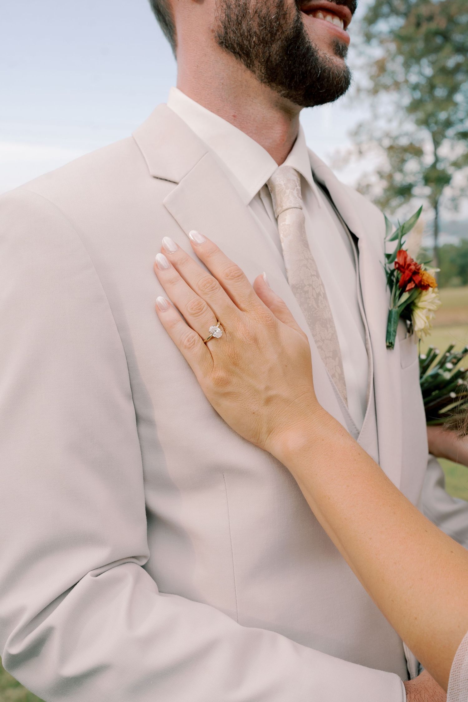 Close-up shot of a hand wearing a wedding ring against a beige suit jacket with a red boutonniere.