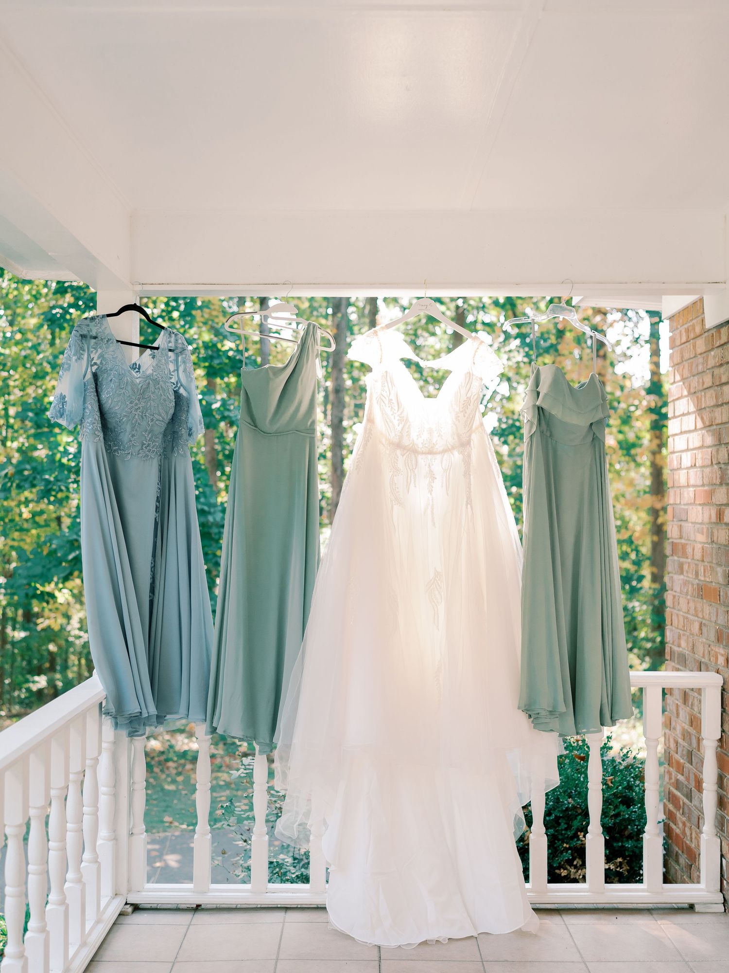Wedding dress and sage green bridesmaid dresses hanging on a white porch railing with greenery in background.