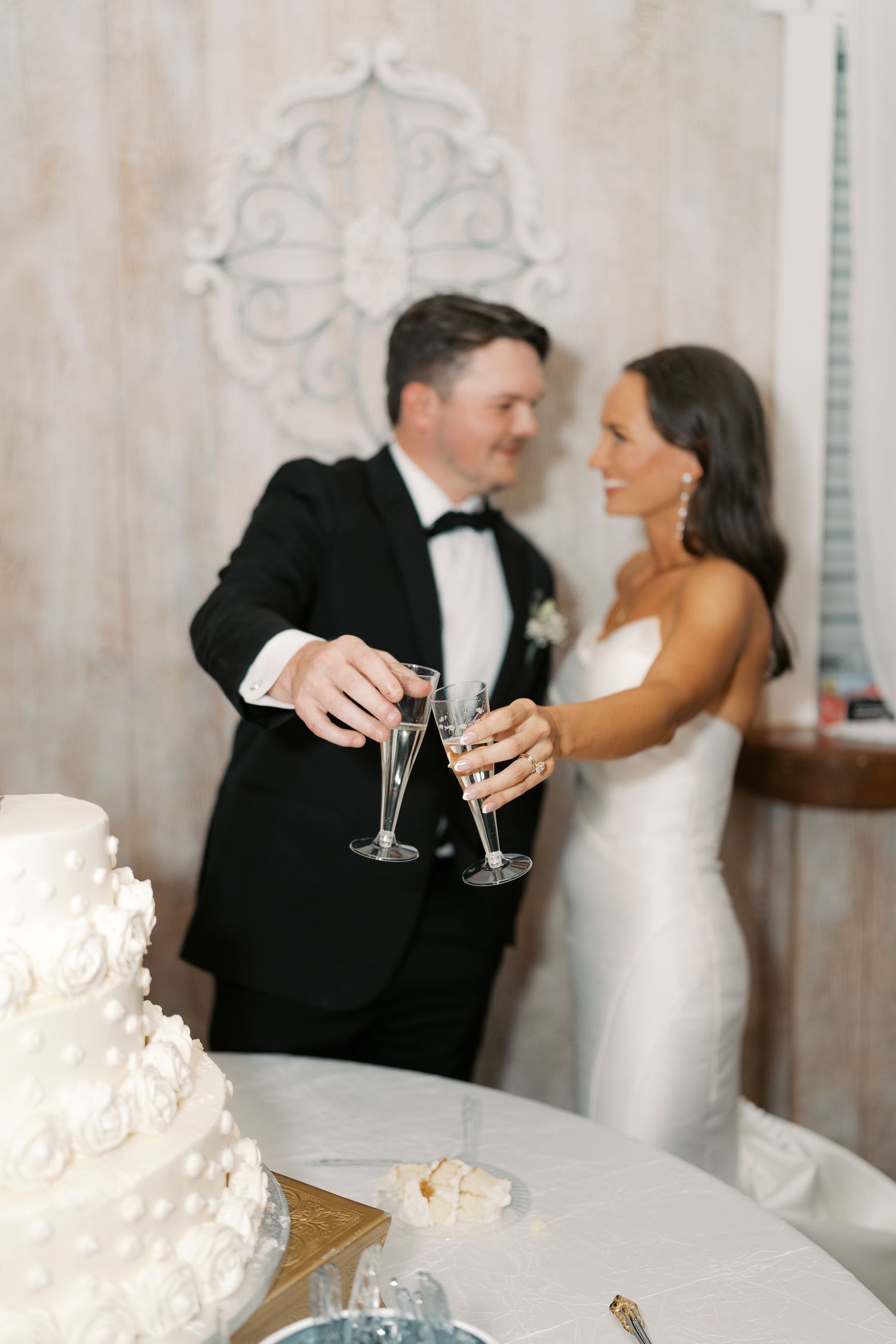 A wedding couple toasts with champagne glasses next to an elegant white tiered wedding cake.