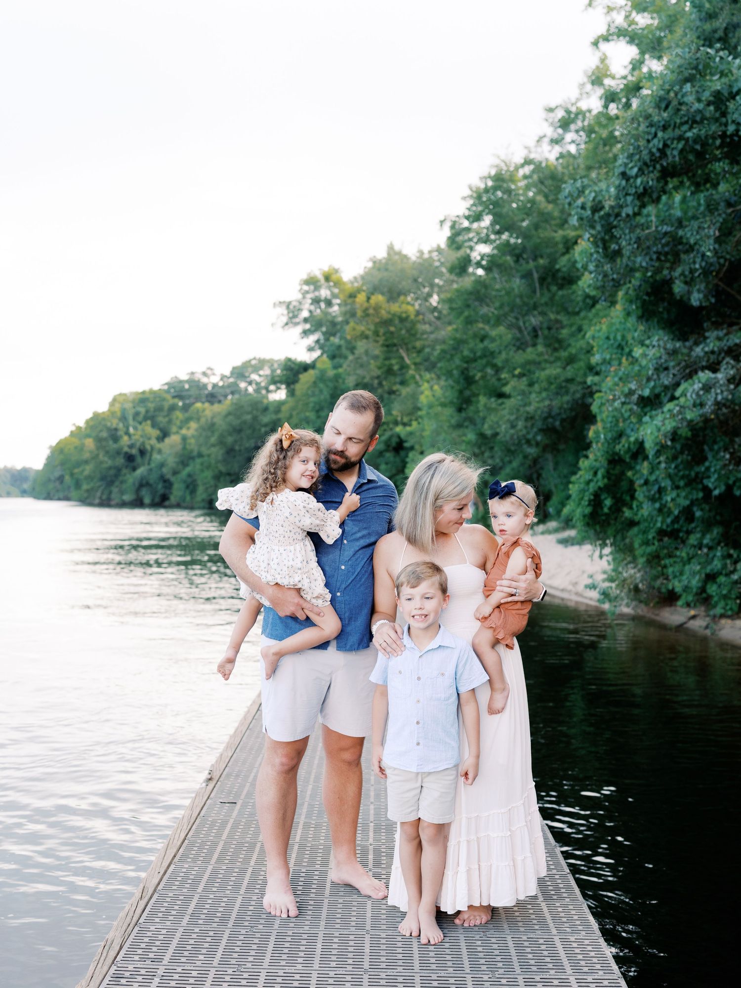 Family portrait on wooden dock by lake surrounded by lush green trees.