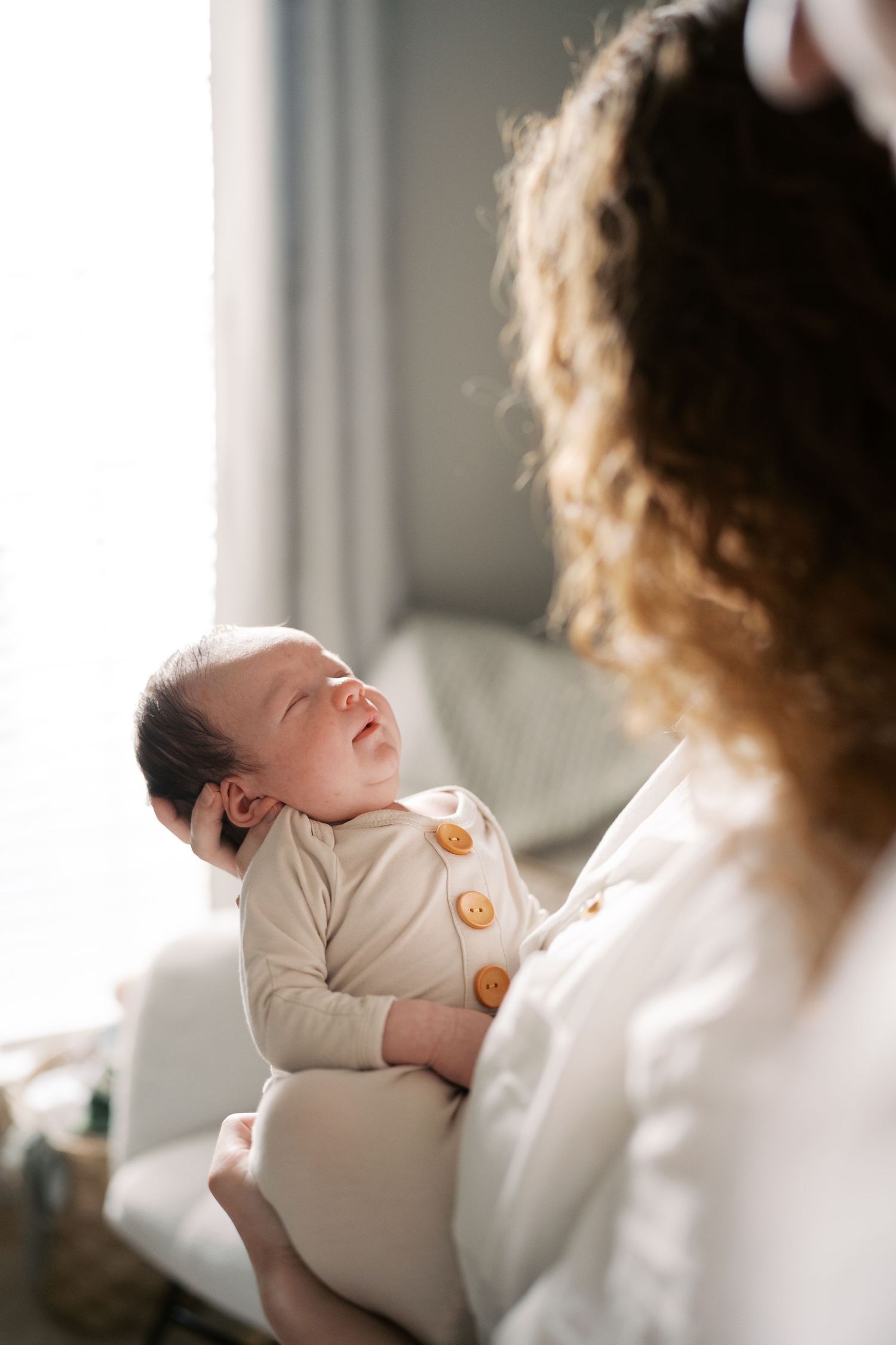A tender moment captured in soft window light shows a baby being cradled in white clothing.