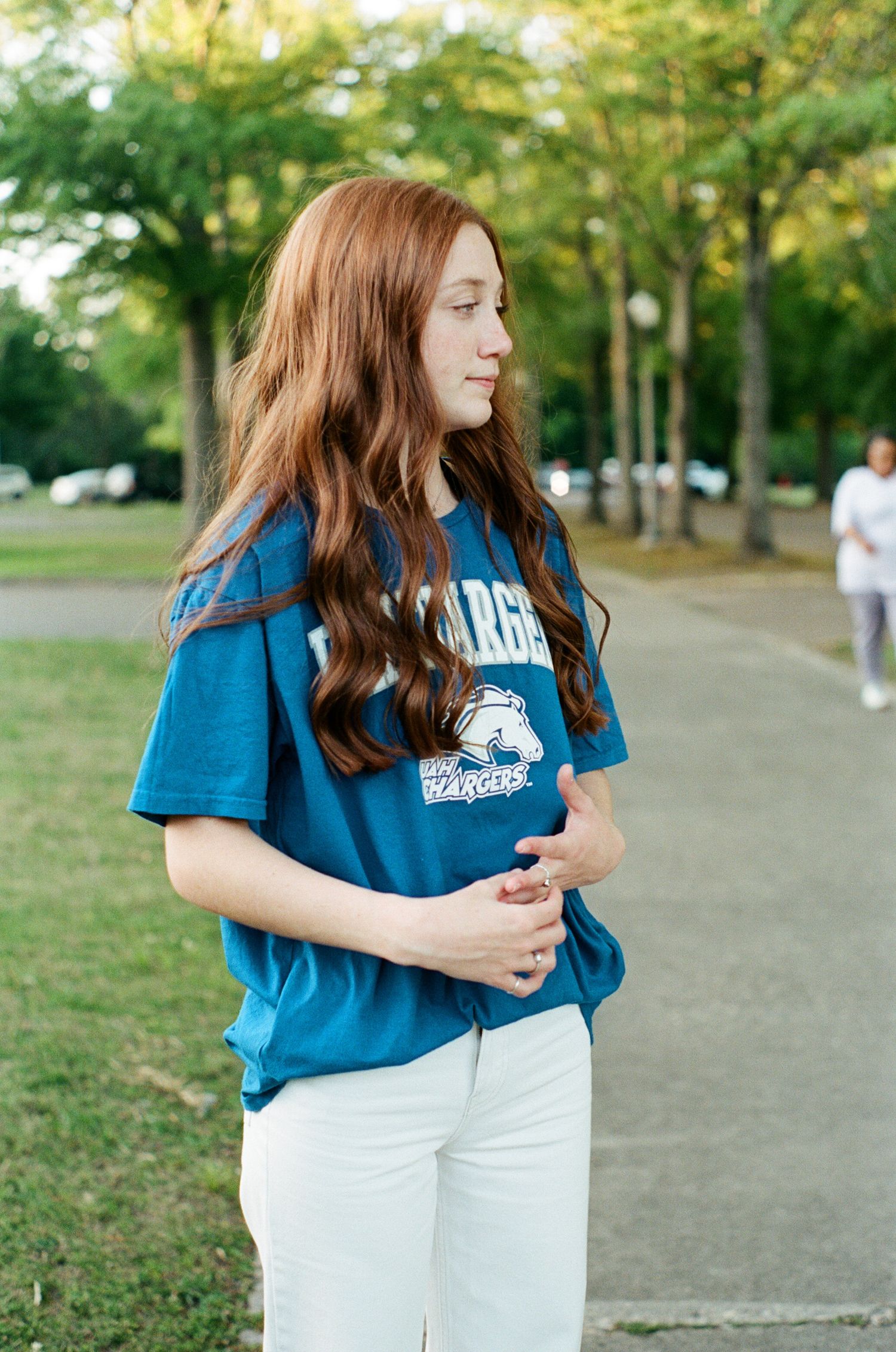 Casual portrait in blue university t-shirt and white pants on tree-lined path.