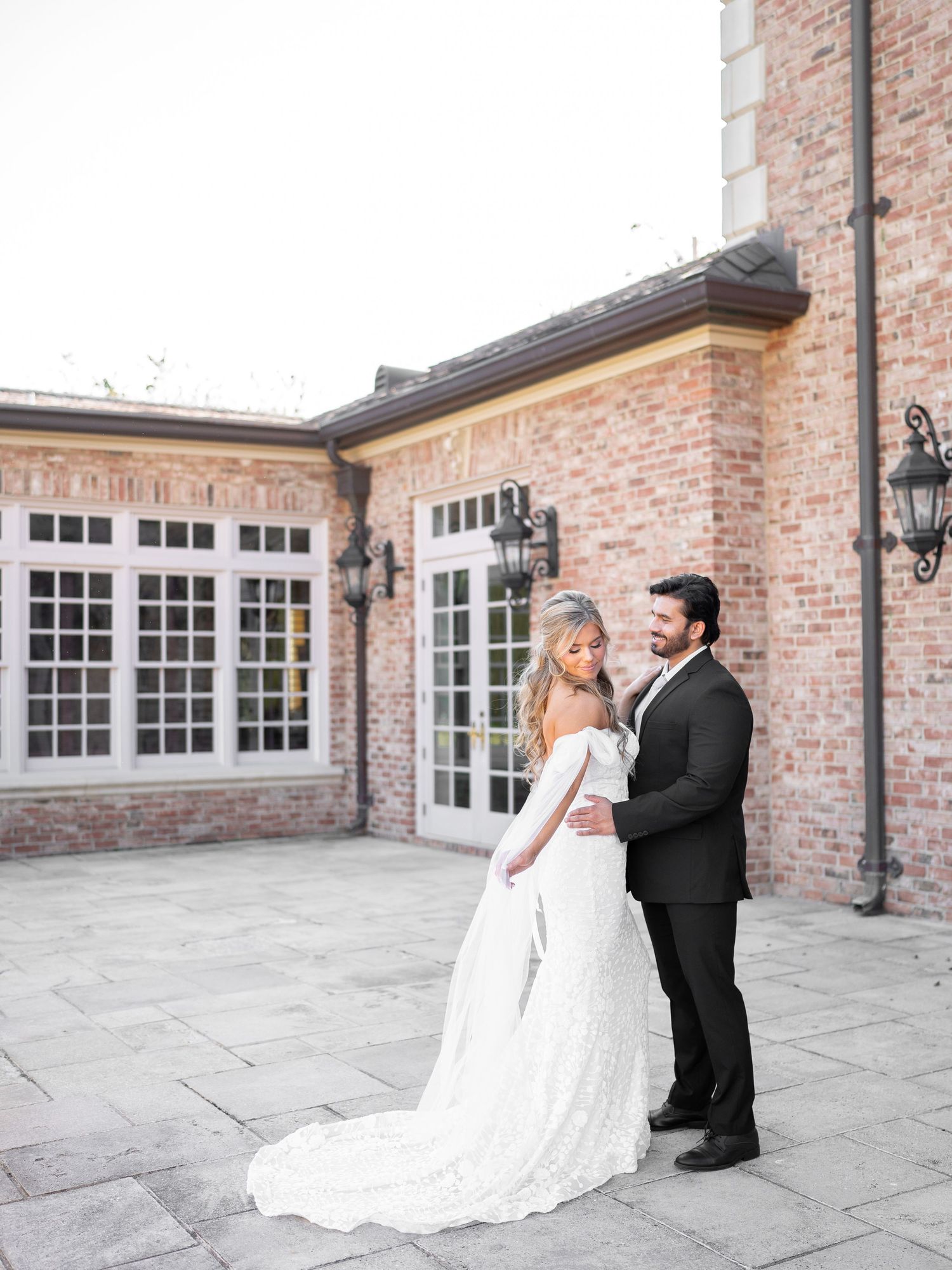 Elegant wedding couple dances on stone patio against brick wall with classic windows.
