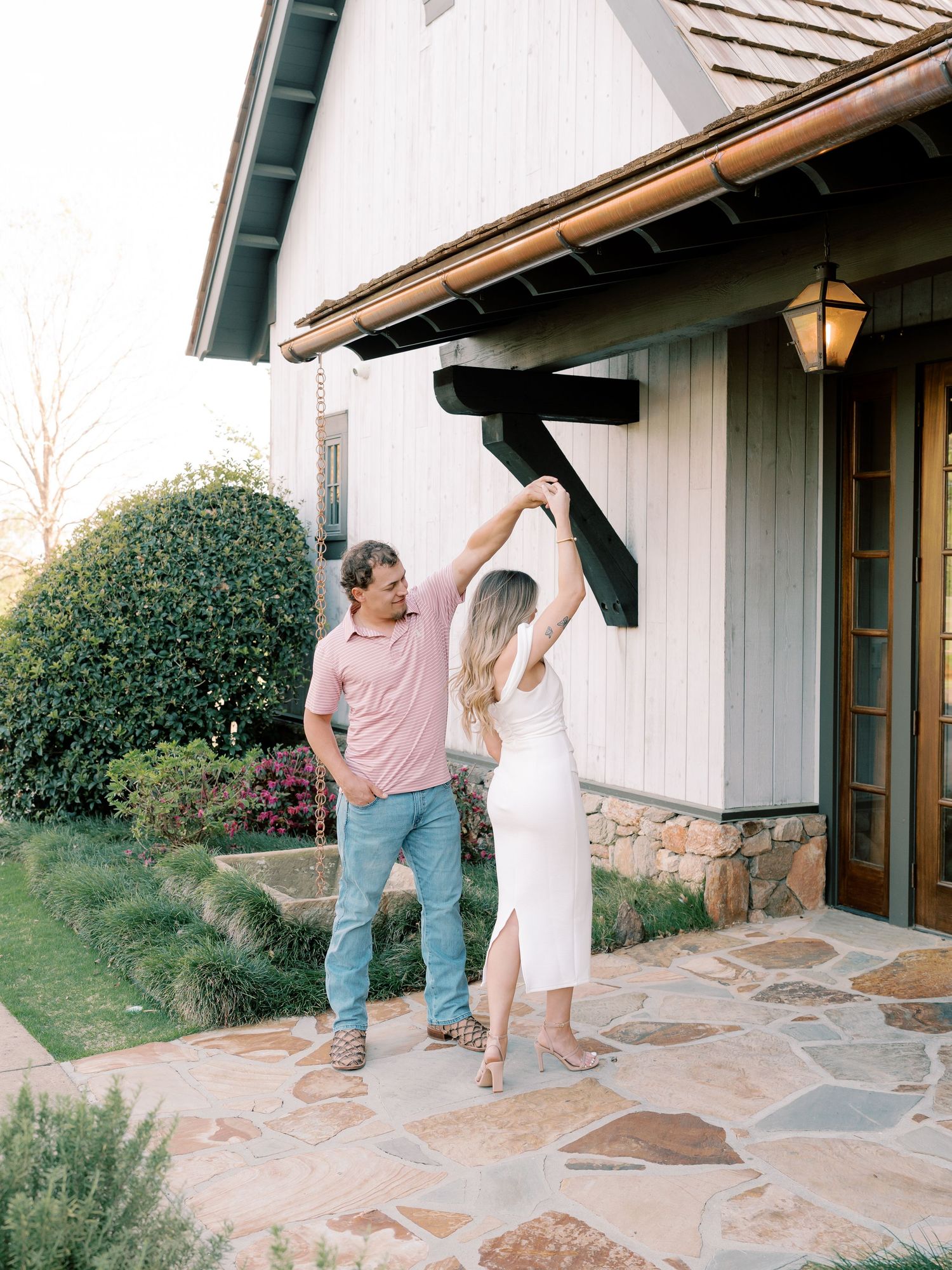 Couple dances and twirls on stone patio outside white building with rustic trim.