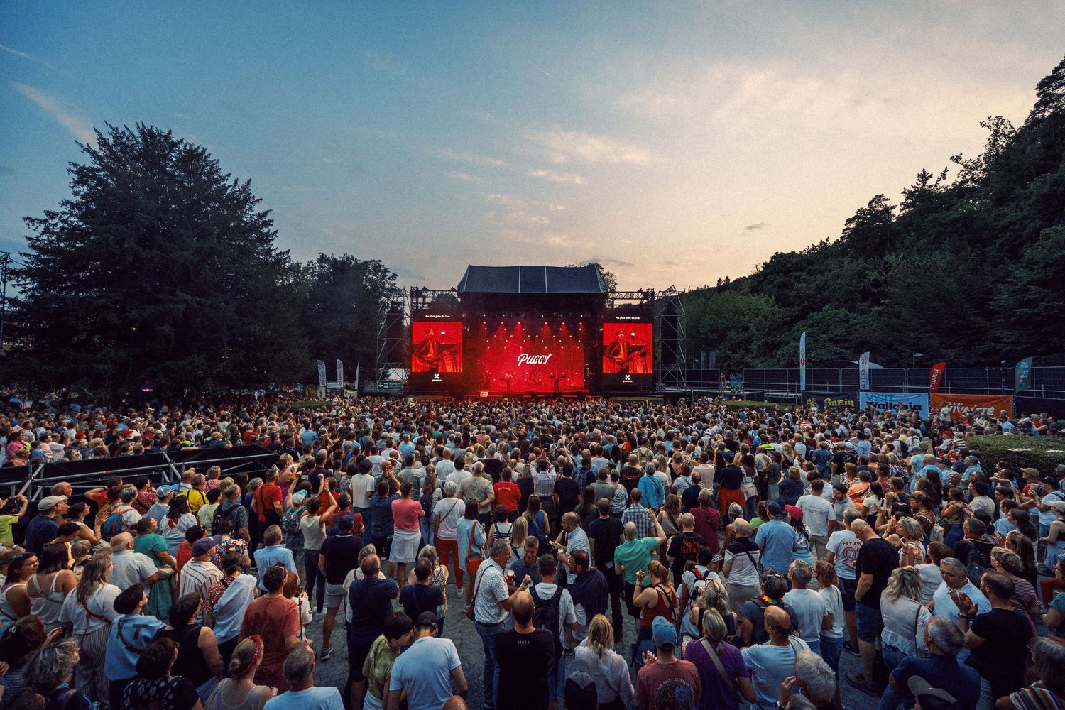 Large outdoor concert crowd at dusk with illuminated red stage surrounded by trees.