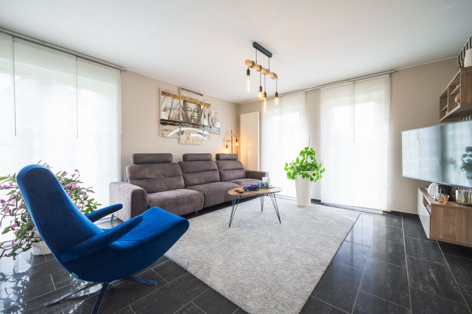 Modern living room with grey sectional sofa, blue accent chair, large windows, and elegant gold chandelier.
