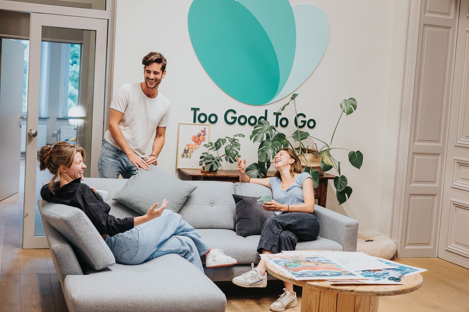 Three colleagues having a casual conversation in a bright office lounge with a Too Good To Go logo on the wall.