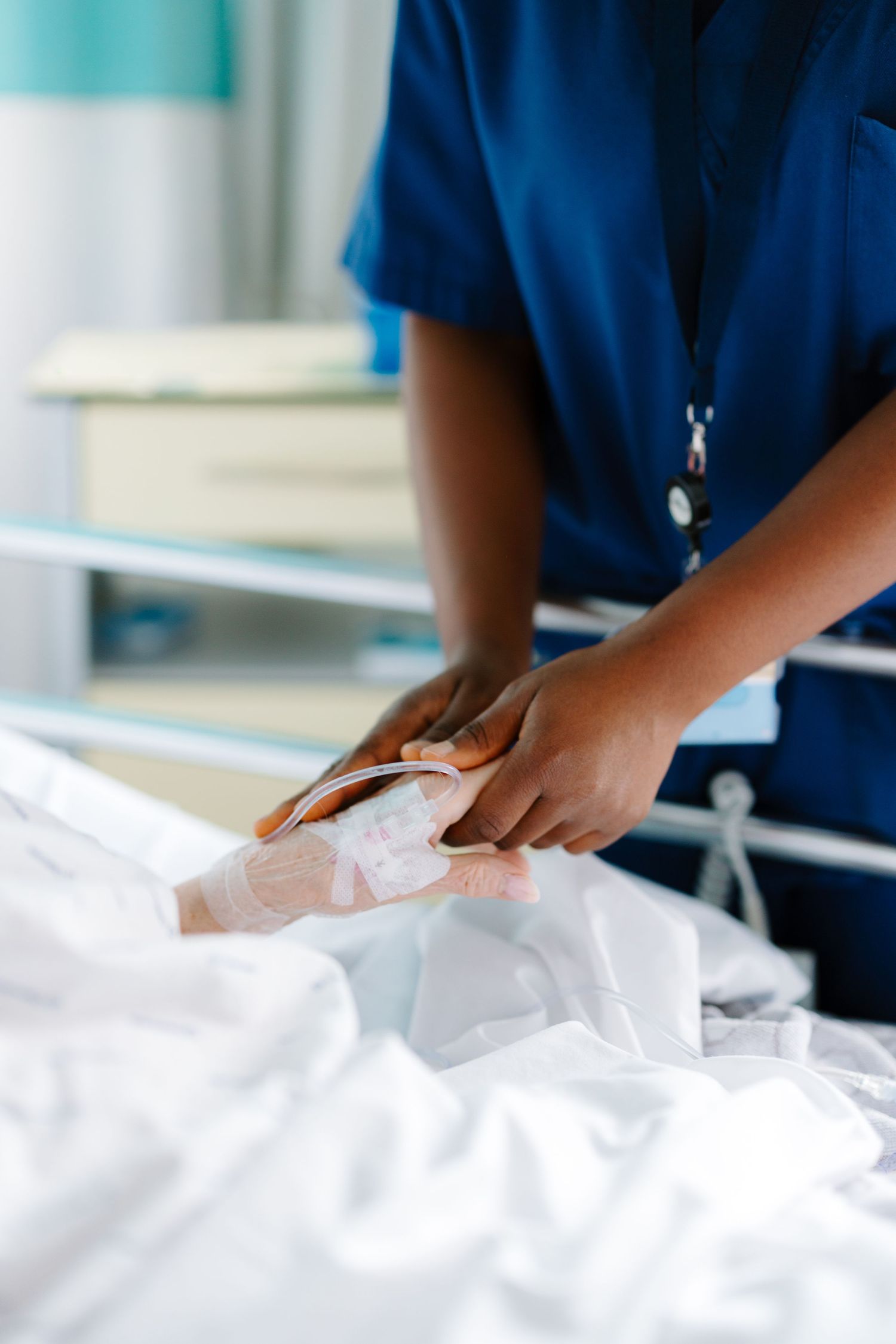 Nurse in blue scrubs checking IV line on patient's hand in hospital bed.