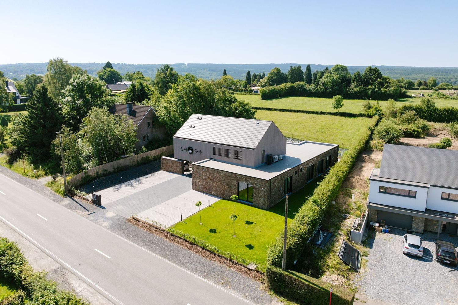 Aerial view of a modern stone house with green lawn, adjacent fields, and scenic countryside views on a quiet road.