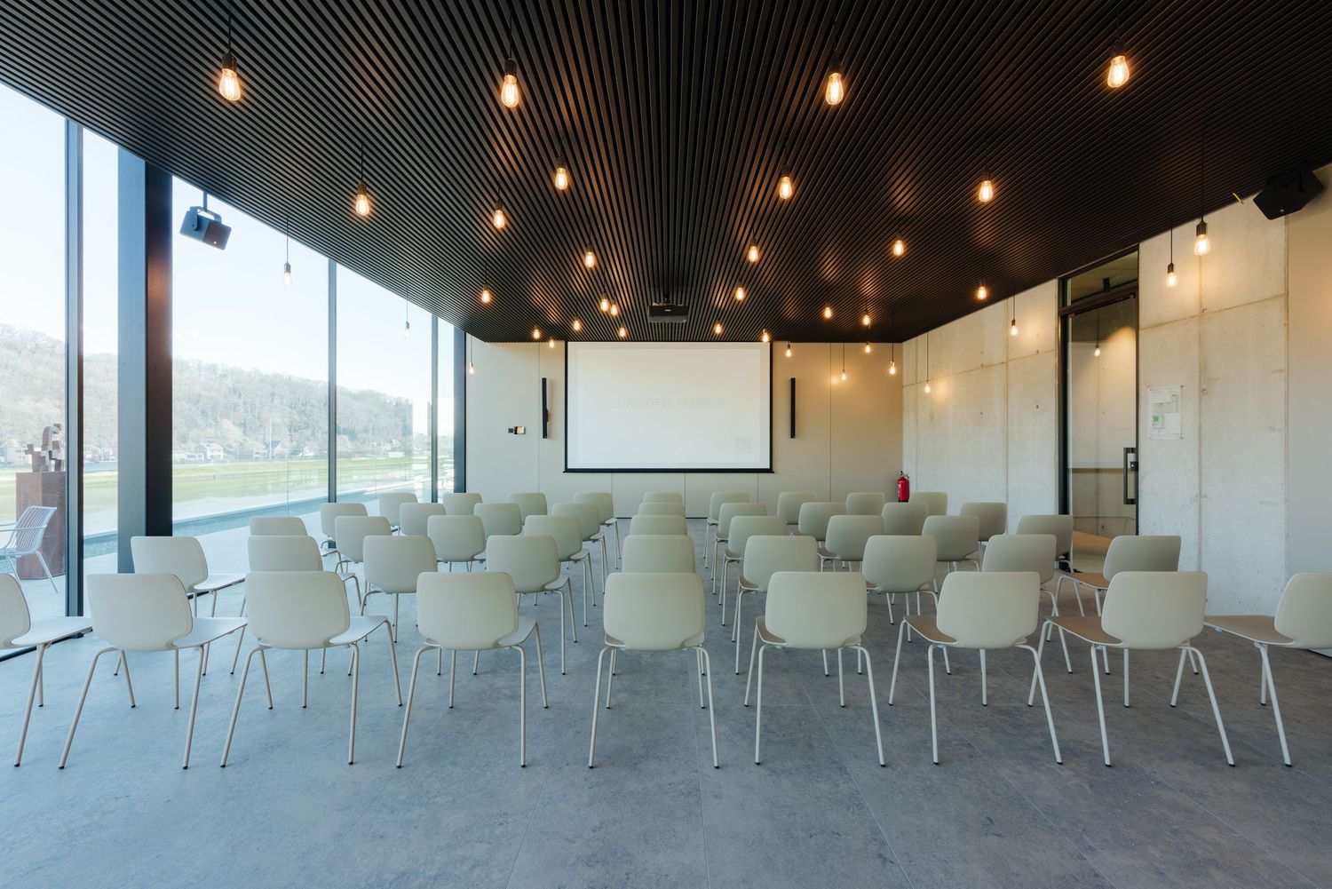 Modern conference room with rows of white chairs facing a projection screen, floor-to-ceiling windows, and pendant lighting.