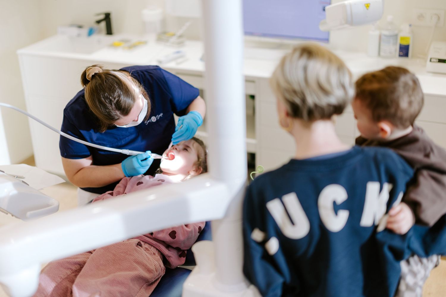 Dentist treating young patient in dental chair while two children watch in modern clinic.