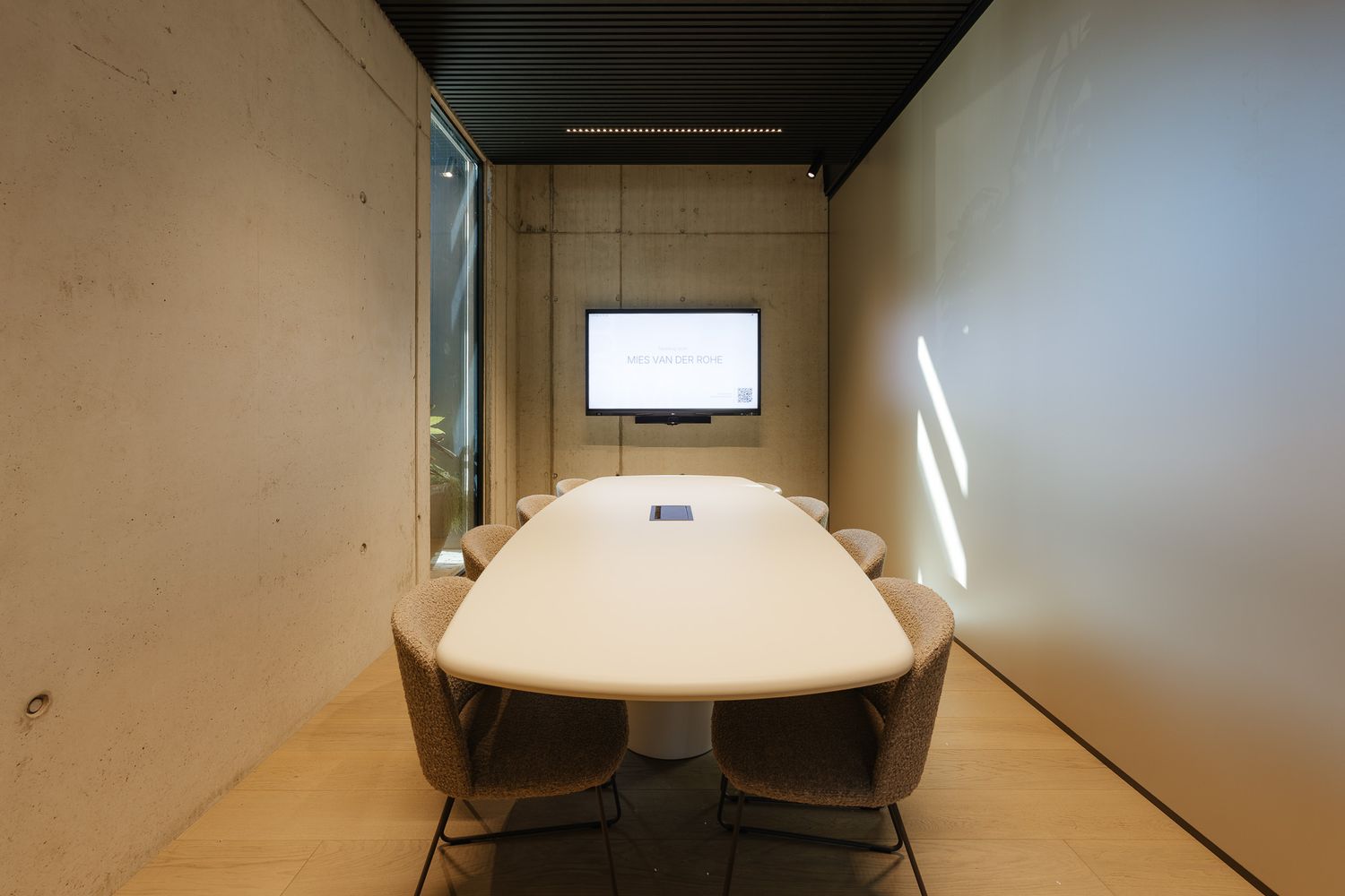 Modern small conference room with oval white table, beige chairs, wall-mounted screen, and concrete walls.
