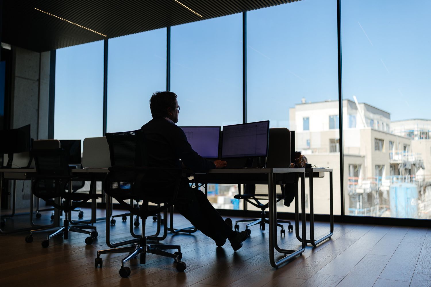 Silhouette of businessman working at dual monitors in modern office with panoramic city windows.
