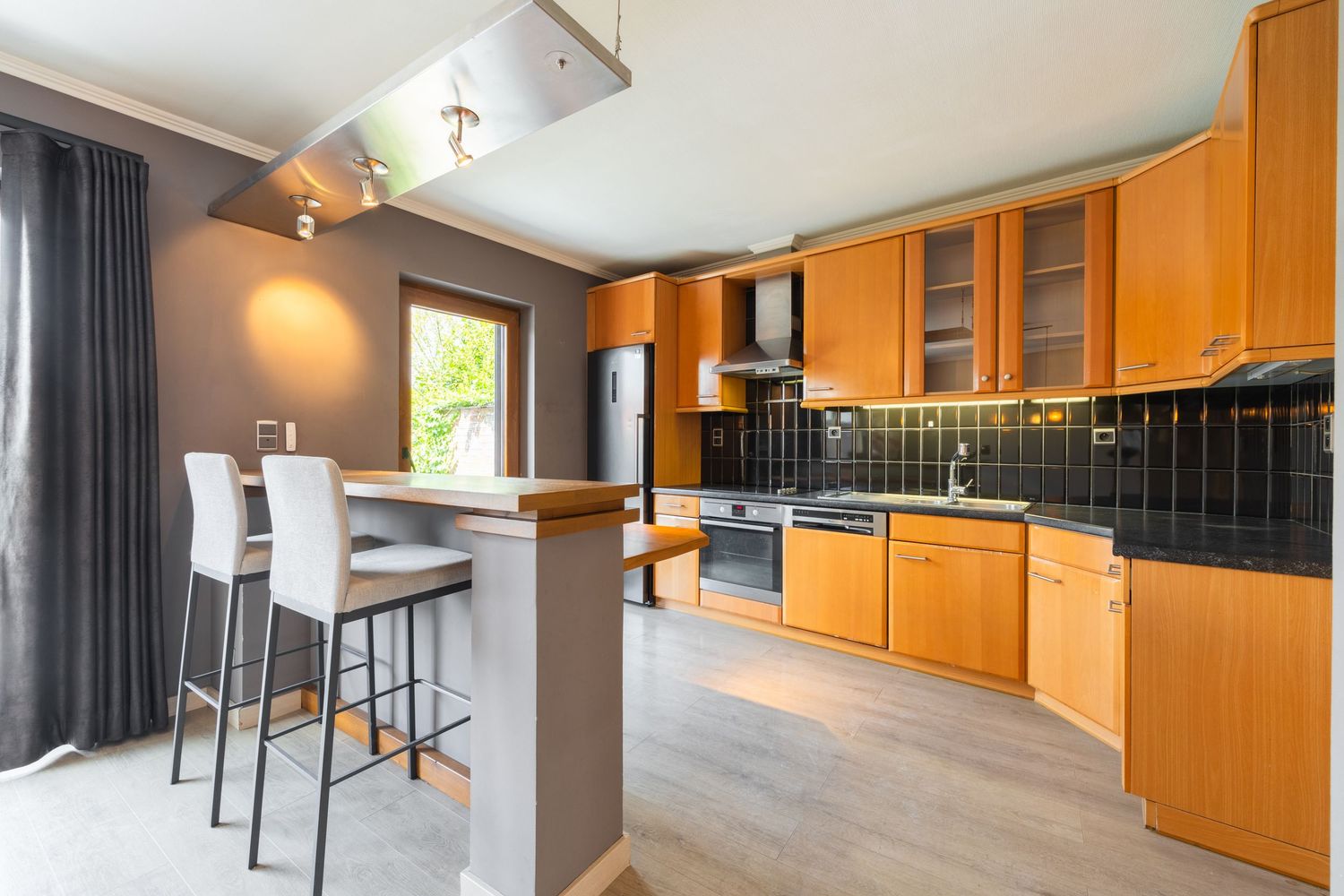 Modern kitchen with wooden cabinets, black tile backsplash, stainless steel appliances, and a breakfast bar with white stools.