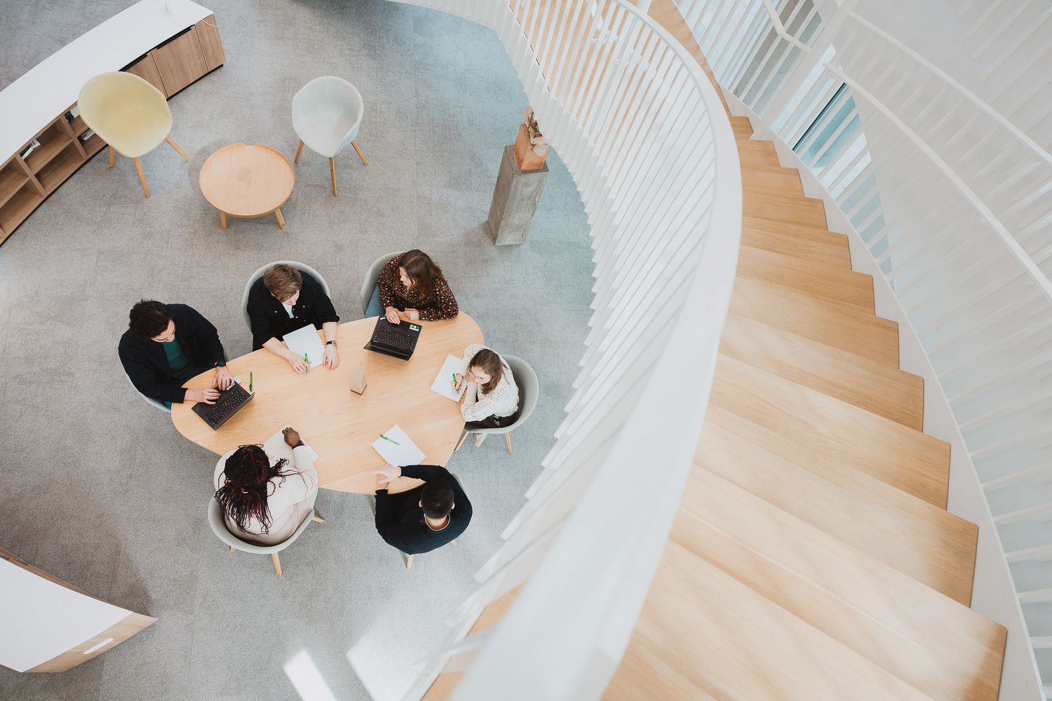 Aerial view of business team meeting around round table near spiral staircase in modern office.