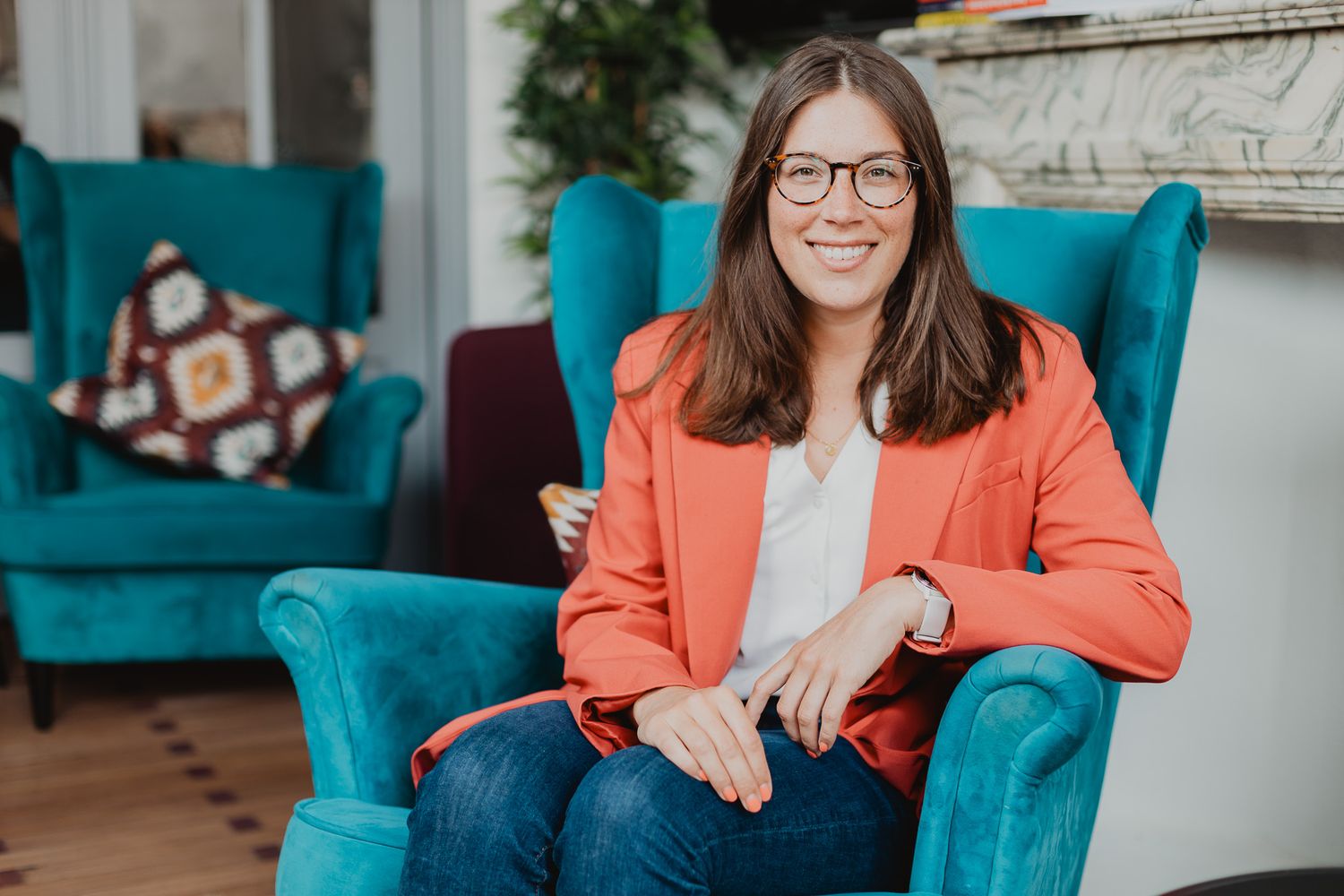 Smiling woman in an orange blazer sitting in a teal velvet armchair in a stylish interior.