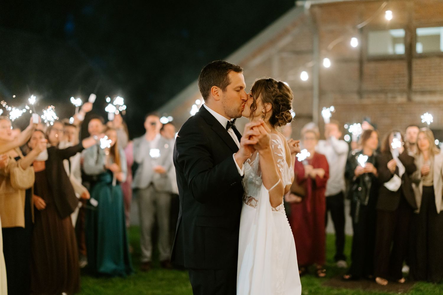 Bride and groom share a kiss surrounded by guests holding sparklers at a romantic outdoor night wedding reception.