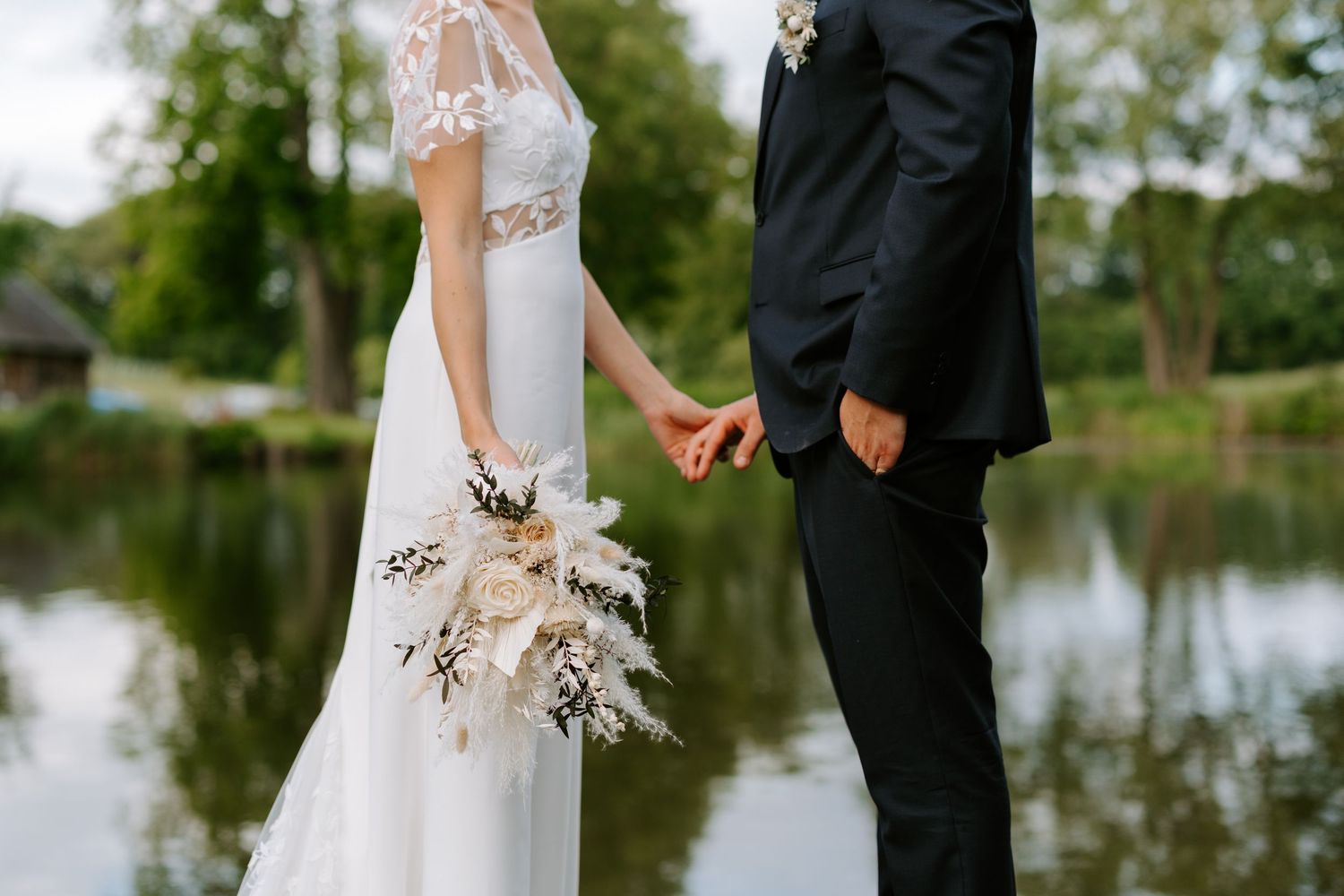 Bride and groom holding hands near a serene lake, bride holding a white boho bouquet in an elegant lace wedding dress.