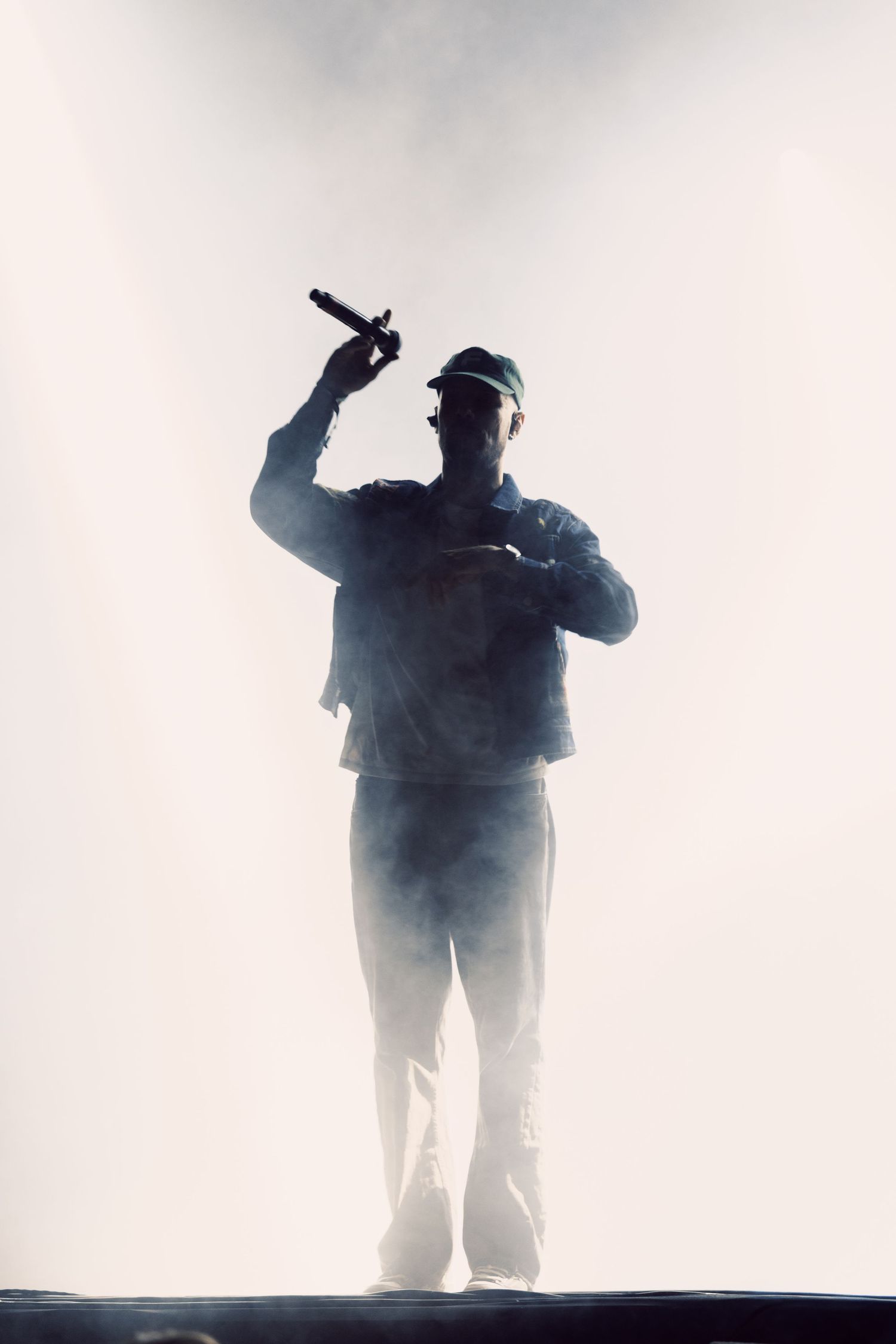 Silhouetted performer holding microphone on stage surrounded by dramatic white smoke and backlighting during a live concert.