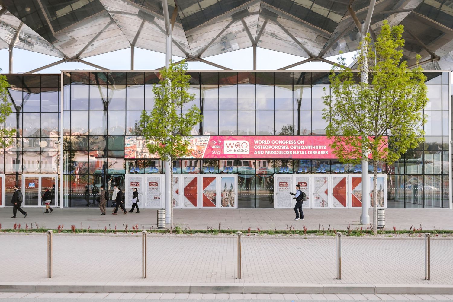 Modern exhibition center facade with red banners, glass windows, and trees lining the entrance plaza.