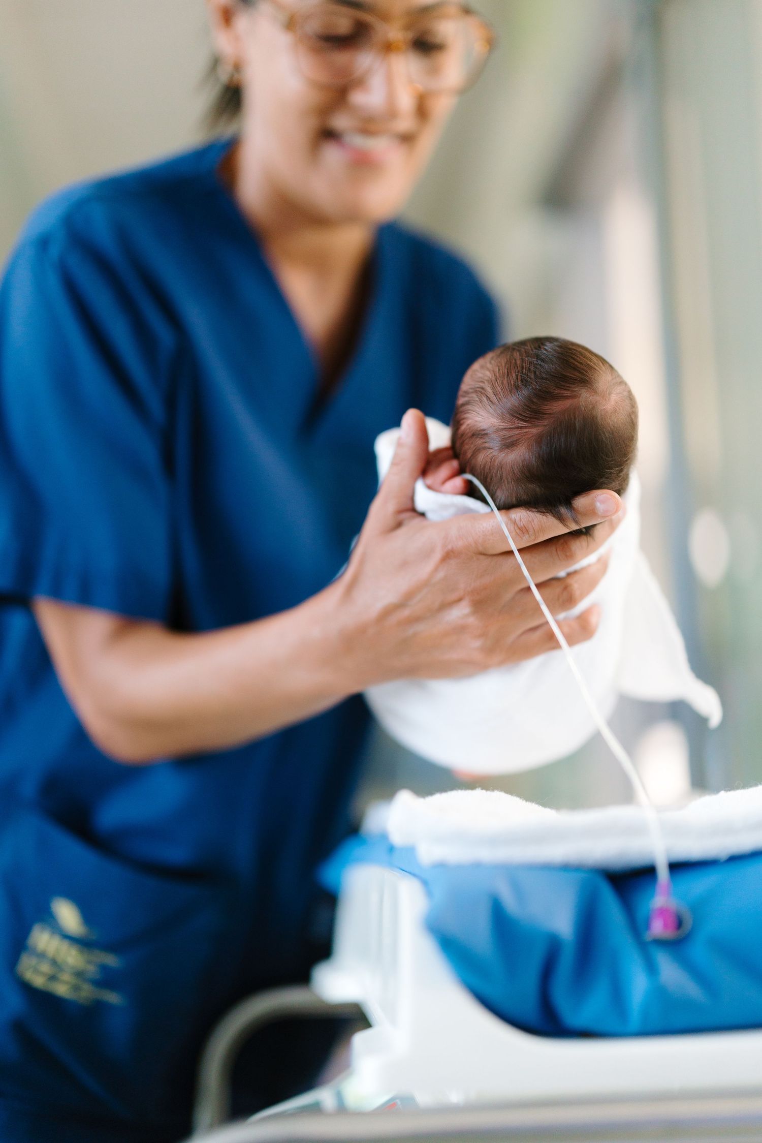 A nurse in blue scrubs gently holds a newborn baby with medical tubes attached in a hospital setting.