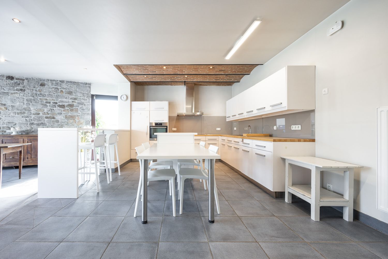 Spacious modern kitchen with white cabinets, stone wall accent, dining table, and large grey floor tiles.