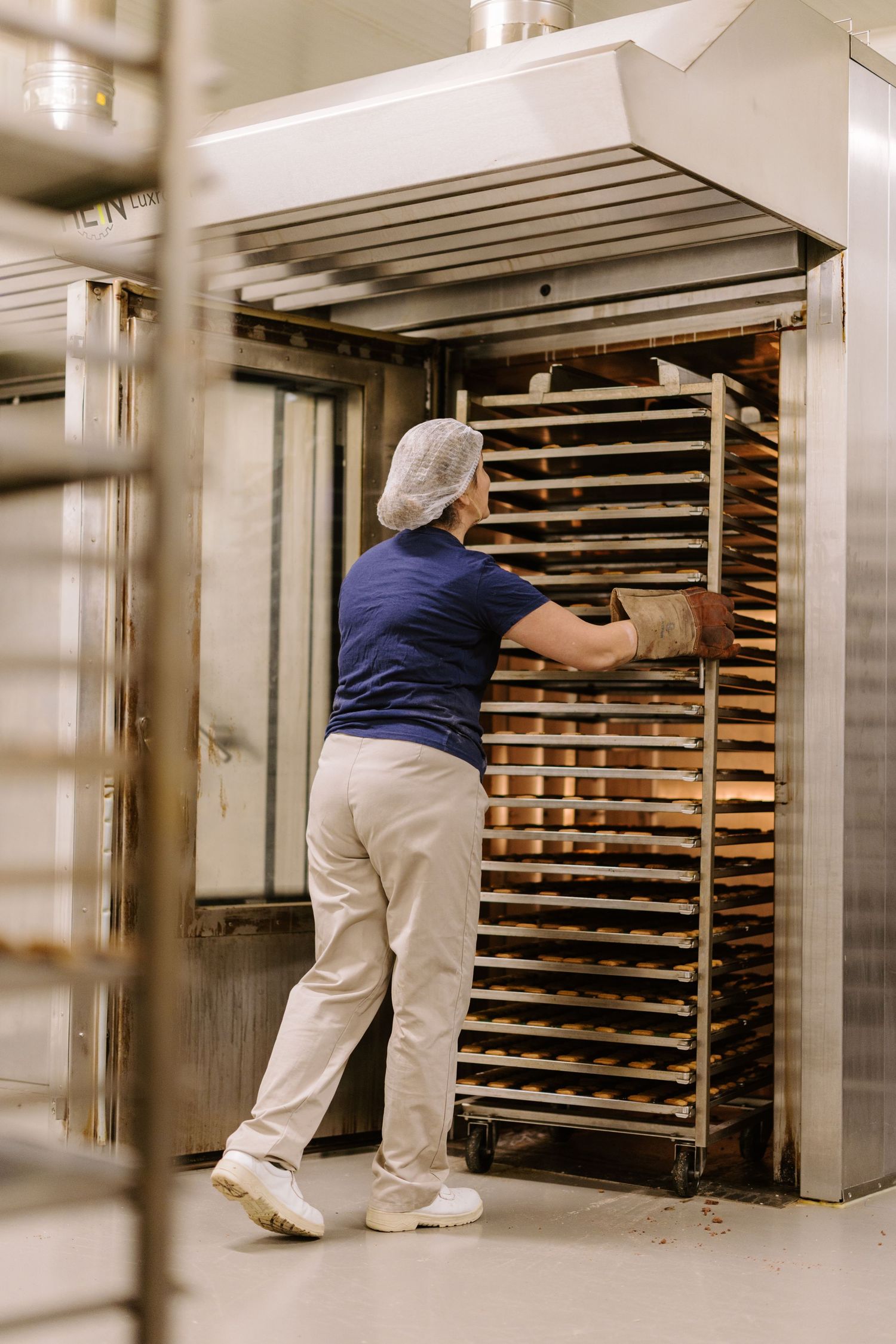 Bakery worker in hairnet loading rack of freshly baked bread into large commercial oven.