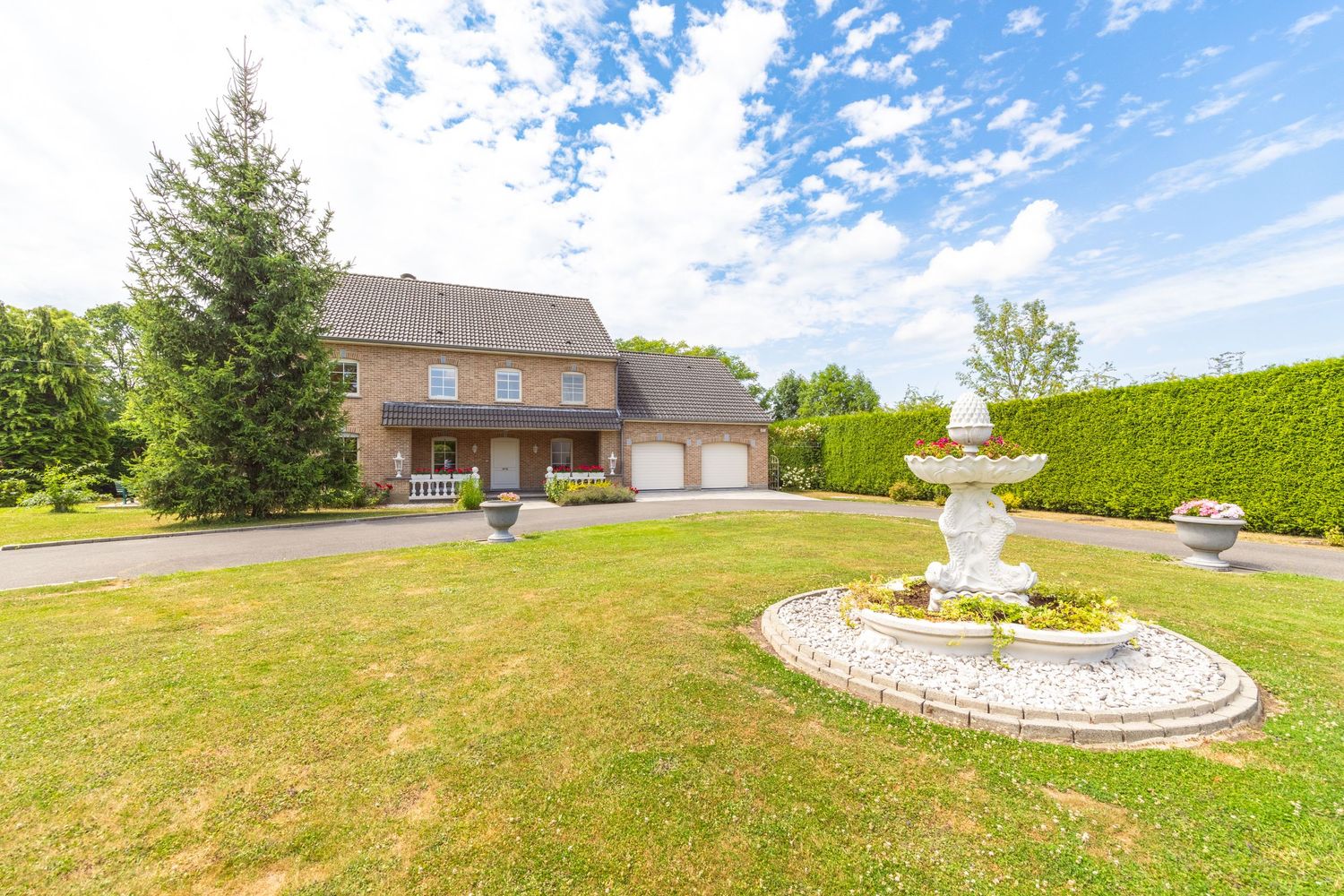 Brick two-story home with attached garage, lush green lawn, ornate fountain centerpiece, tall hedges, and evergreen trees on a sunny day.