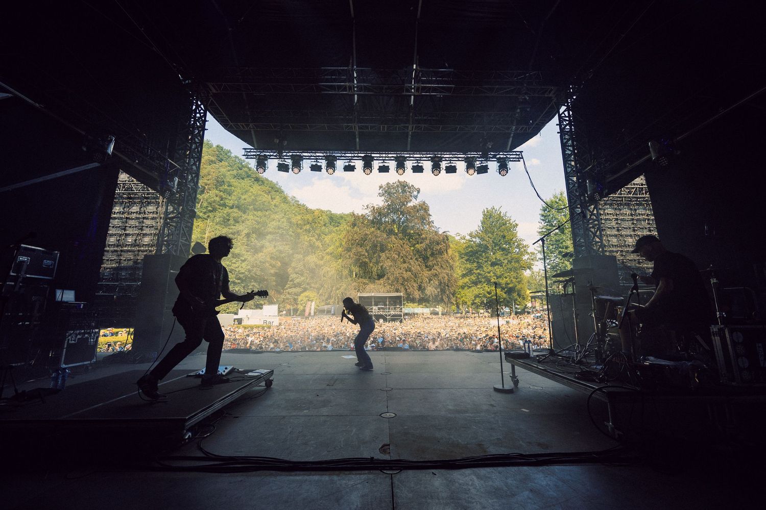 Silhouetted band performing on large outdoor festival stage with massive crowd and green hills in background.
