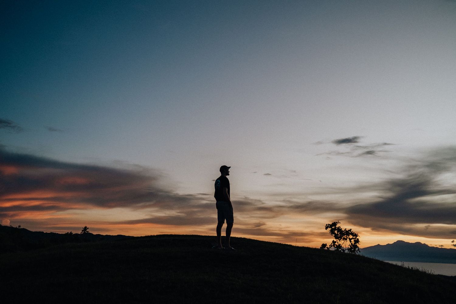 Silhouette of a lone person standing on a hilltop watching a dramatic colorful sunset over the ocean.