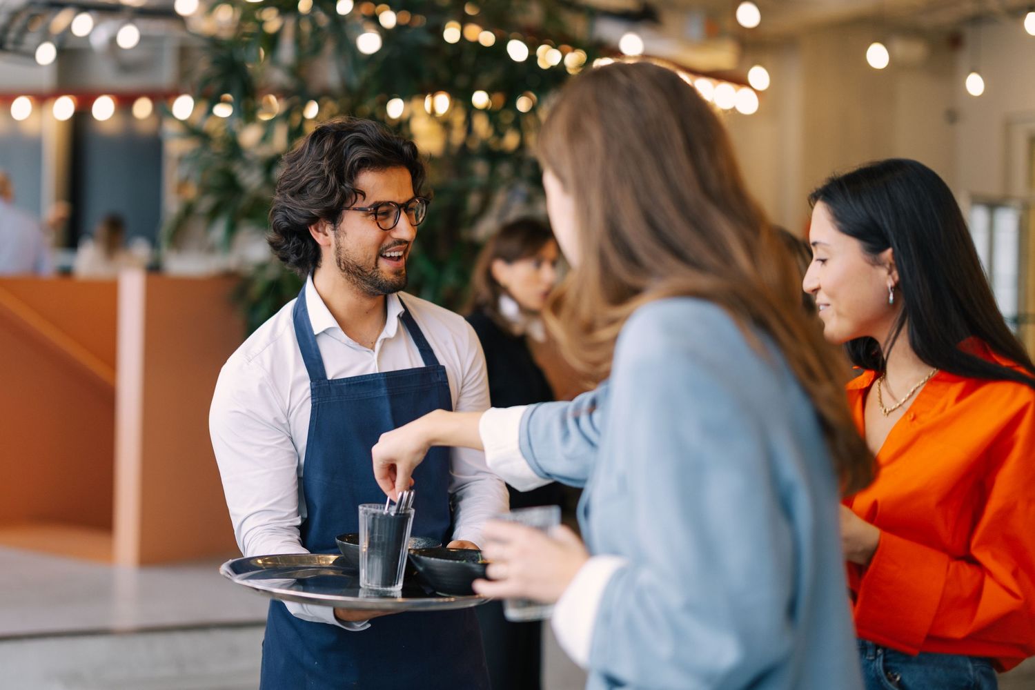 Friendly waiter in apron serving drinks on a tray to smiling female guests in a warmly lit restaurant.
