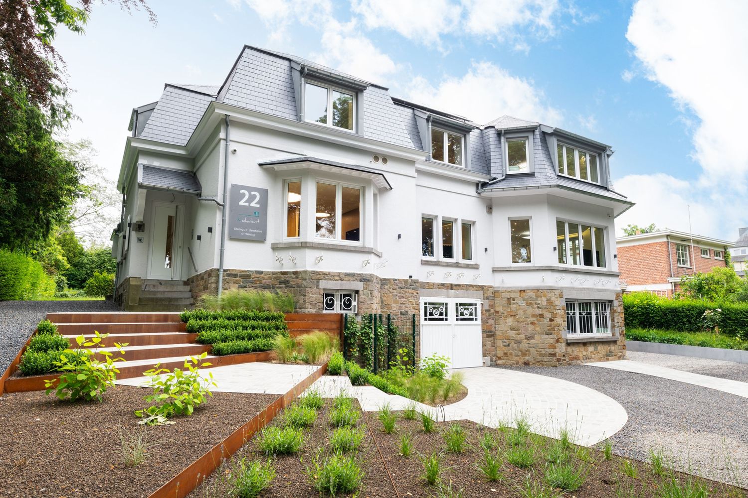 Modern white luxury home with manicured gardens, stone accents, and elegant dormer windows on a sunny day.