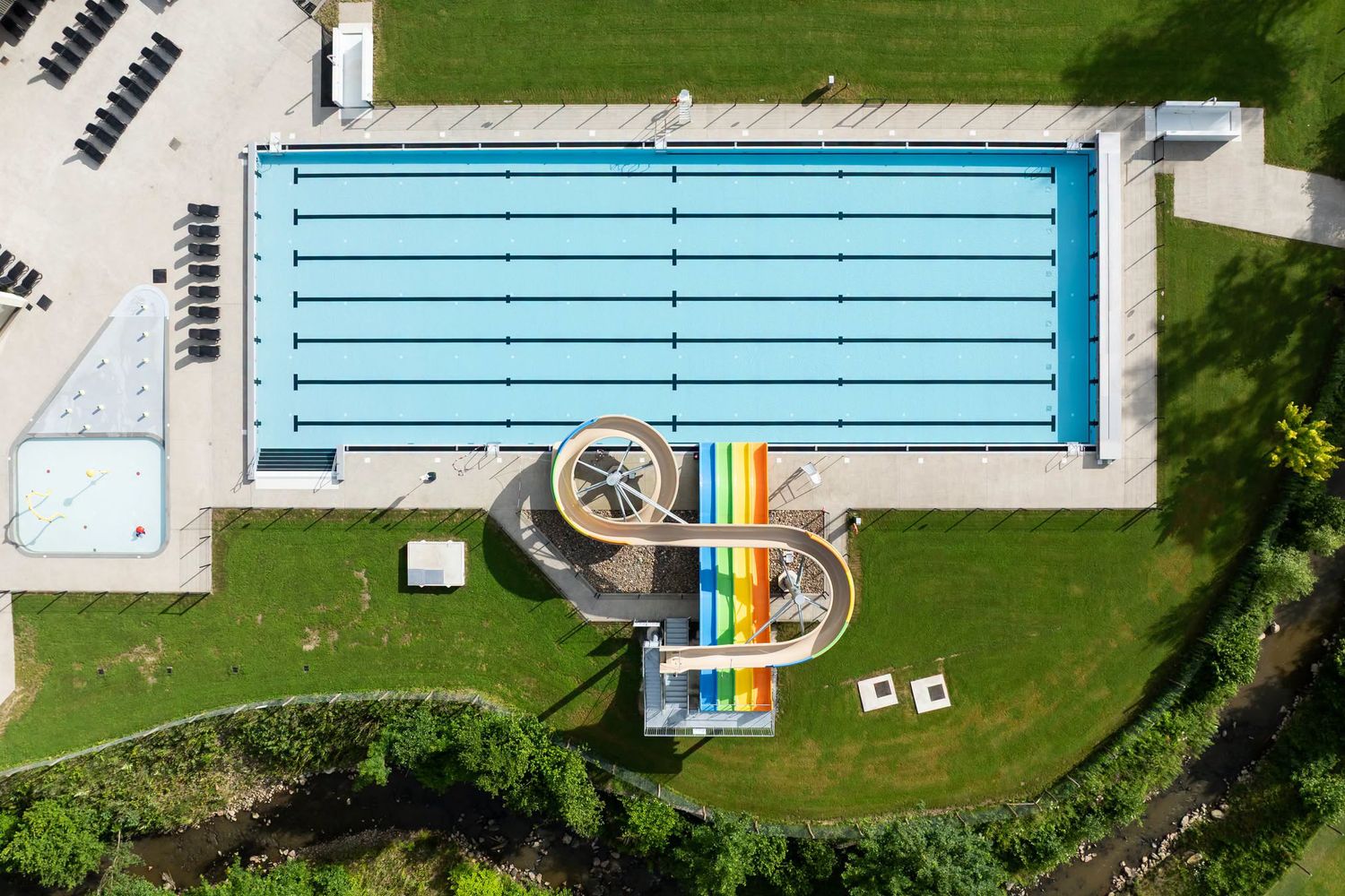 Aerial view of an outdoor swimming pool complex with a large lap pool and colorful water slides on green grass.
