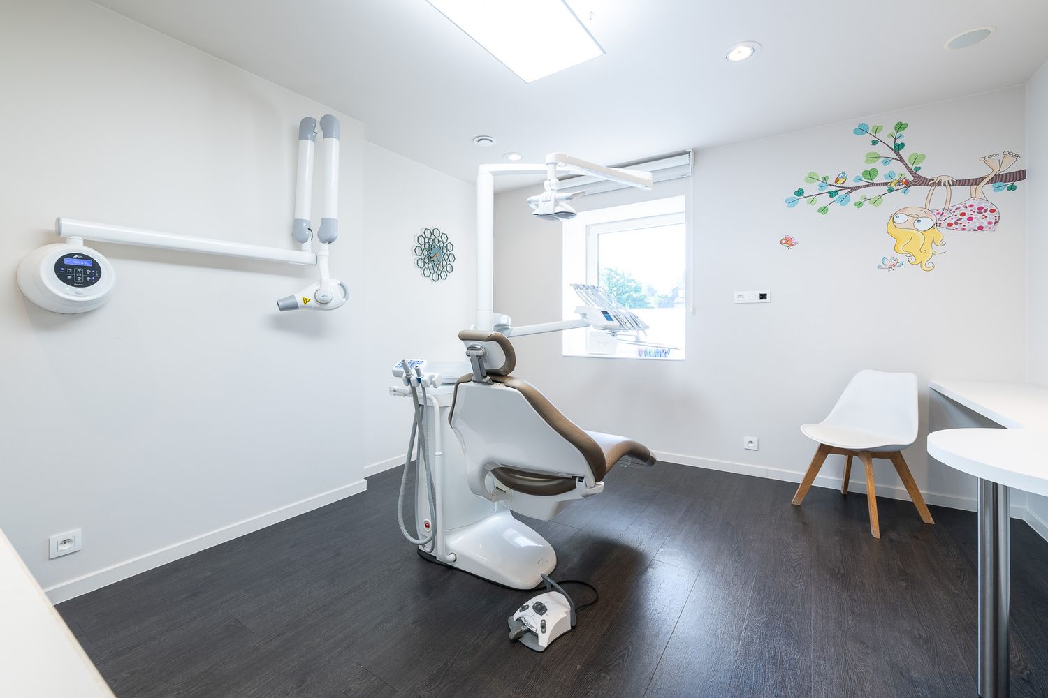 Modern dental treatment room with chair, equipment and colorful tree mural on white wall.