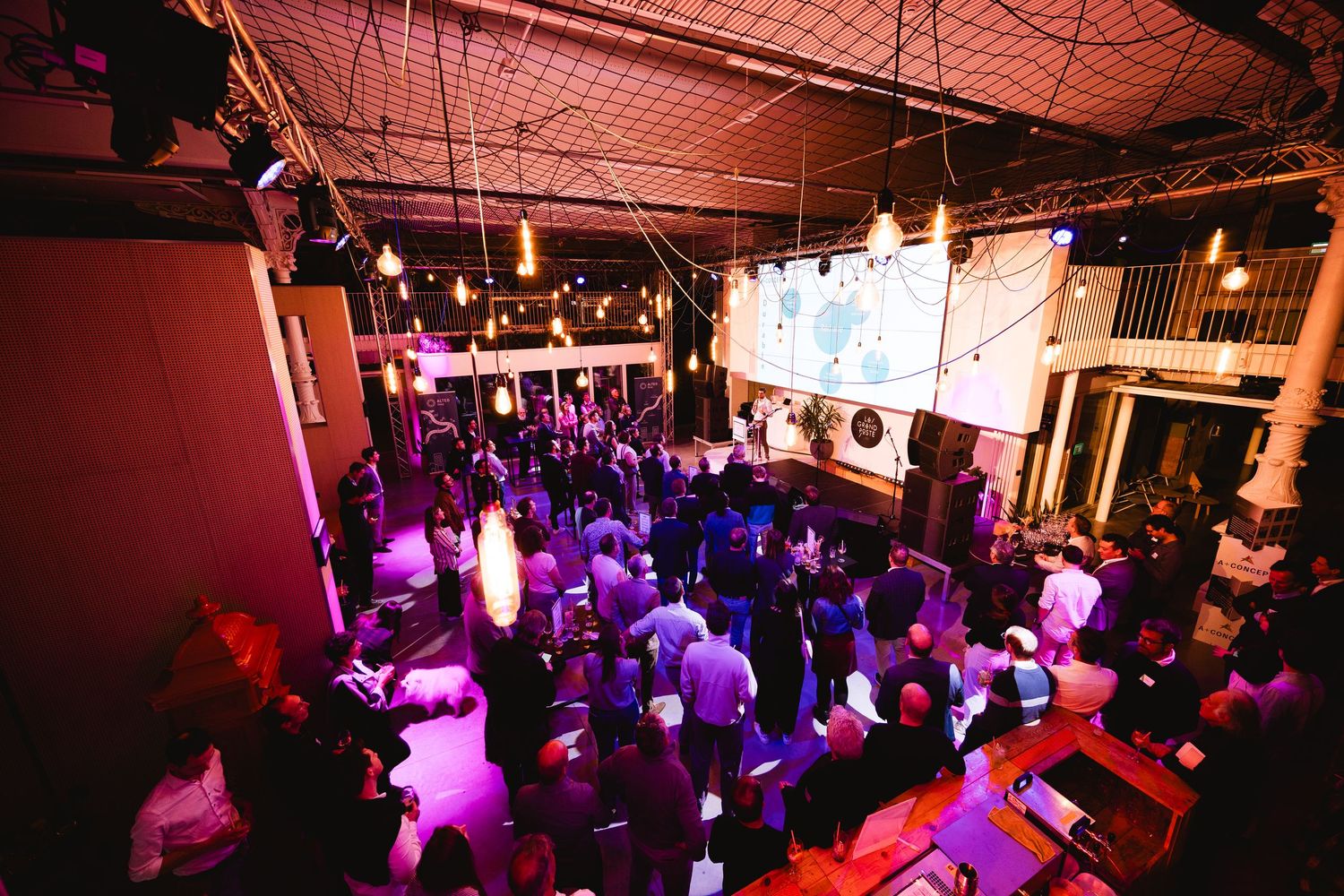 Crowded nightclub event with purple lighting and large projection screens overhead.