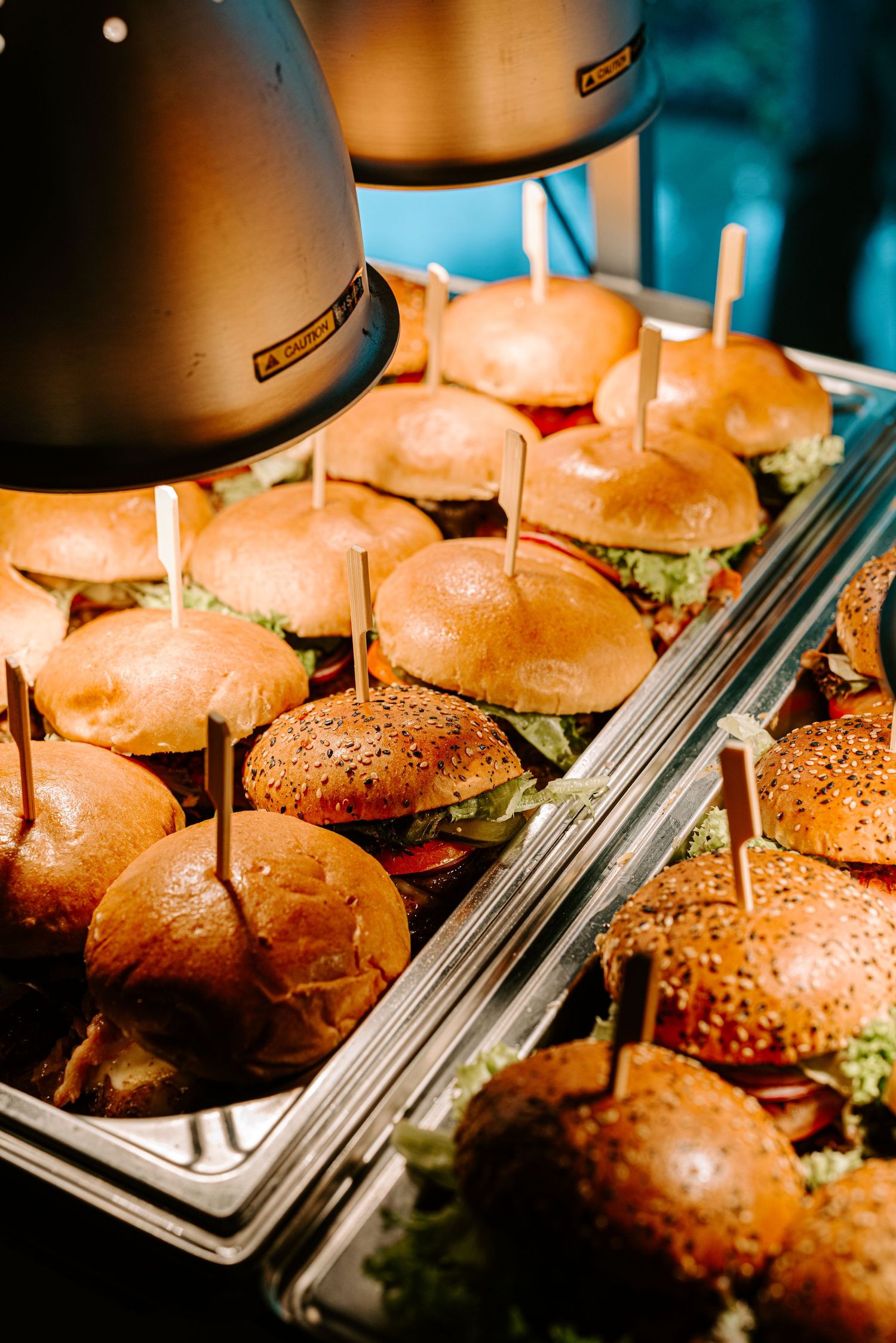 Tray of freshly prepared mini burgers with sesame buns kept warm under heat lamps at a buffet.
