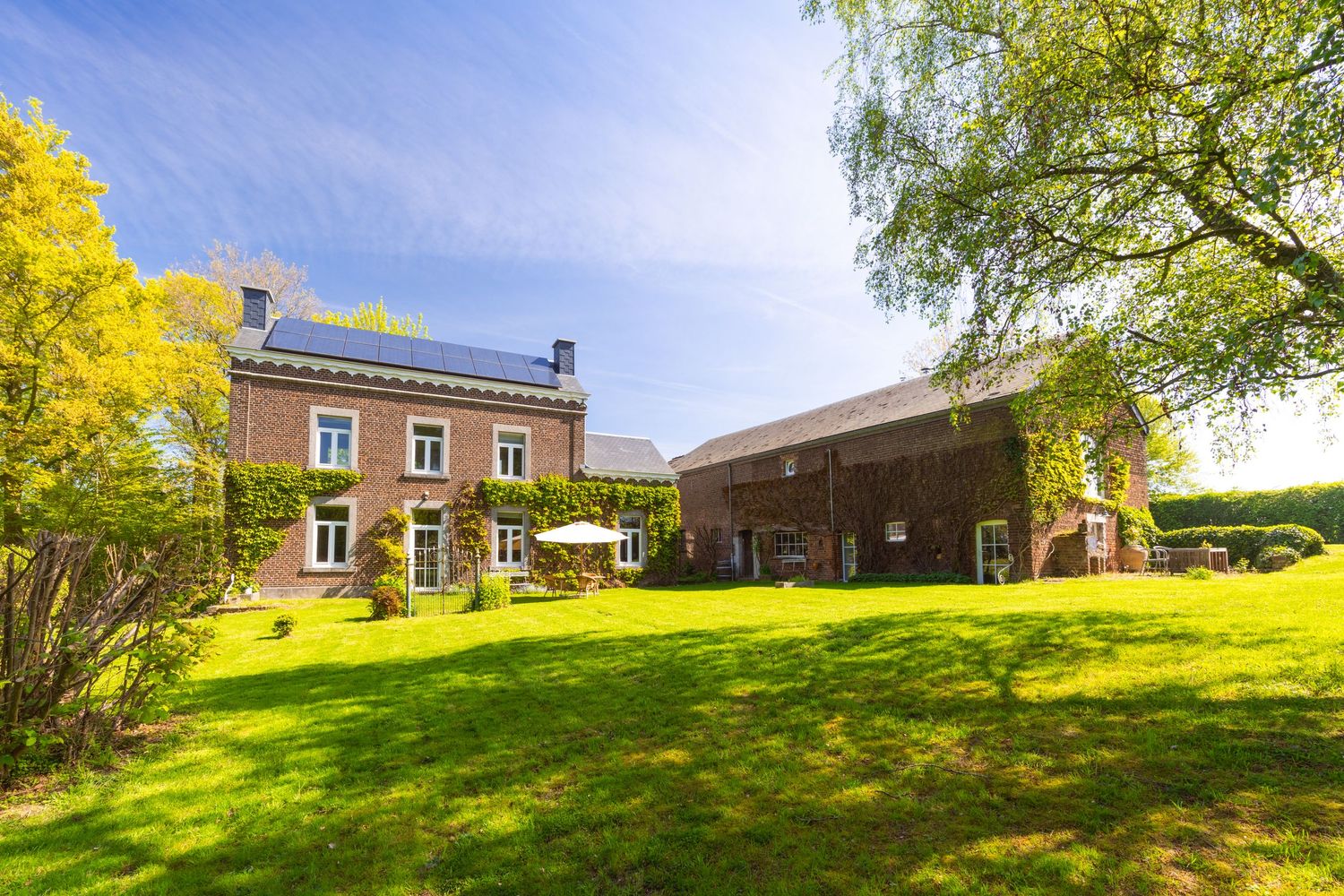 Charming Belgian brick farmhouse with solar panels, lush green lawn, and barn under a bright blue sky.