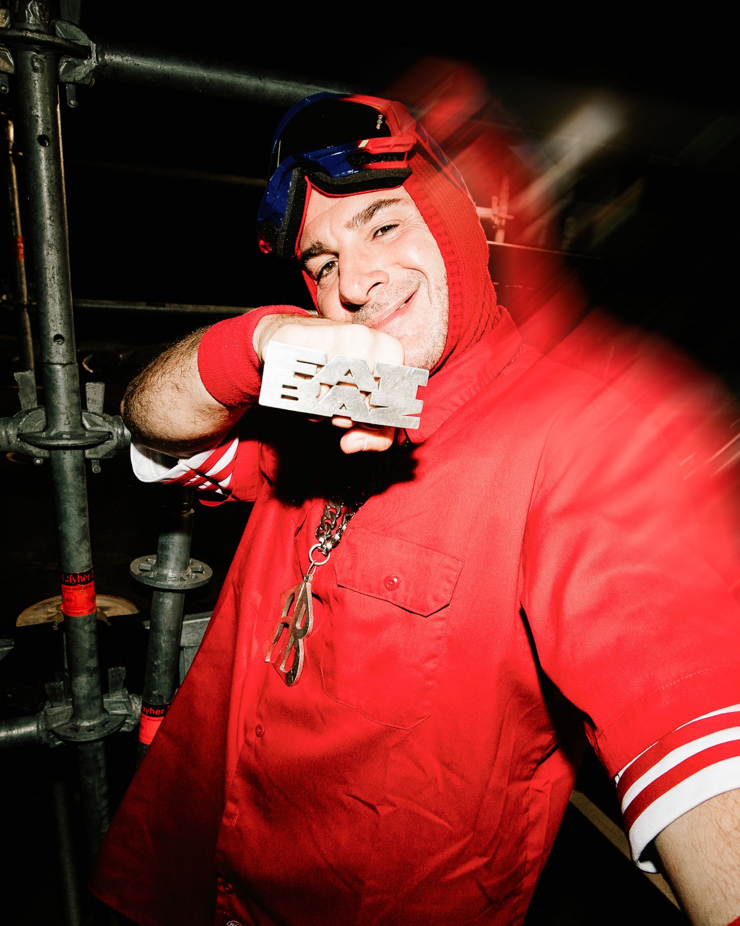 Man in red hoodie with blue goggles smiling and holding a white card in a dark industrial setting.
