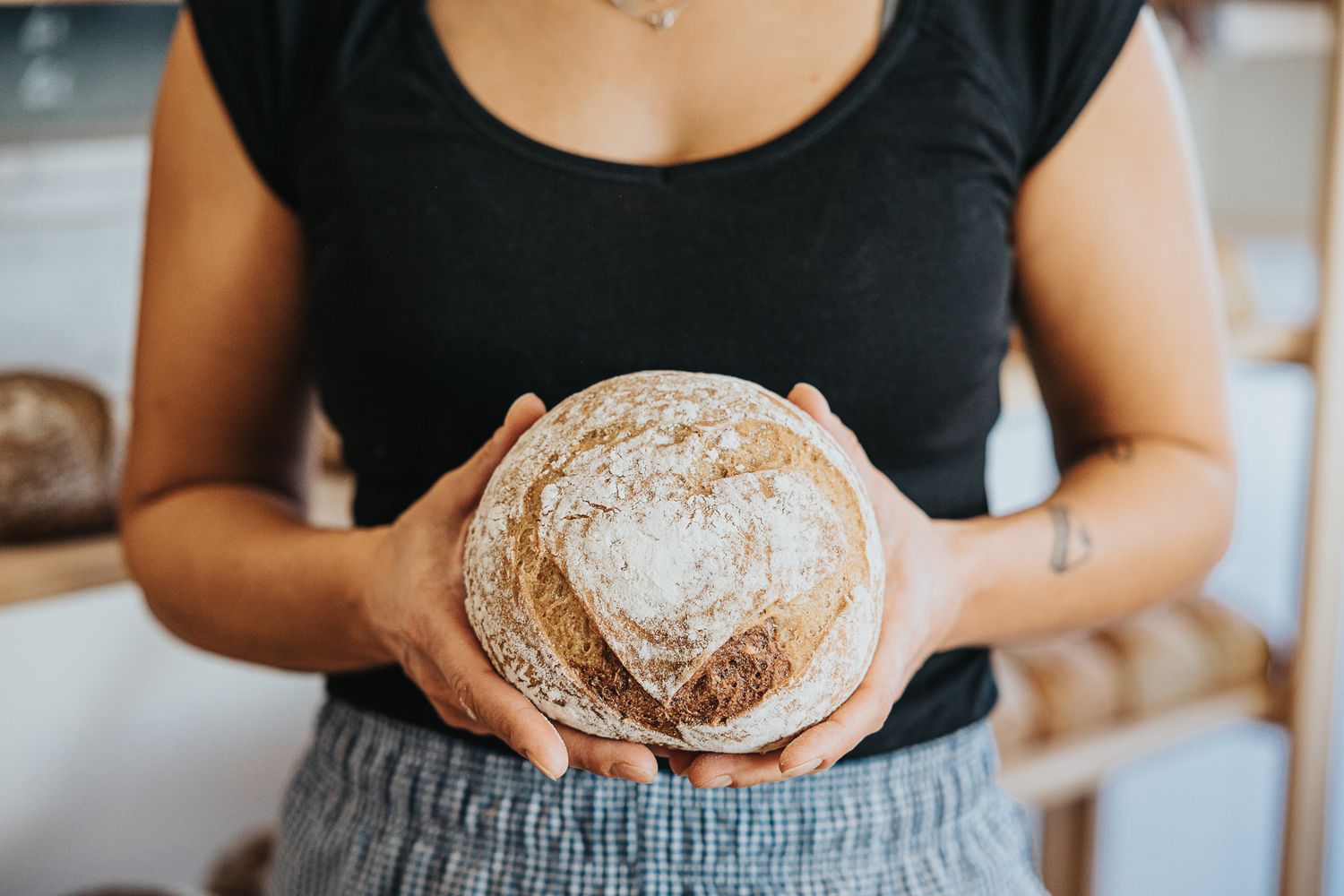 Woman in black top holding freshly baked round sourdough bread loaf in artisan bakery.