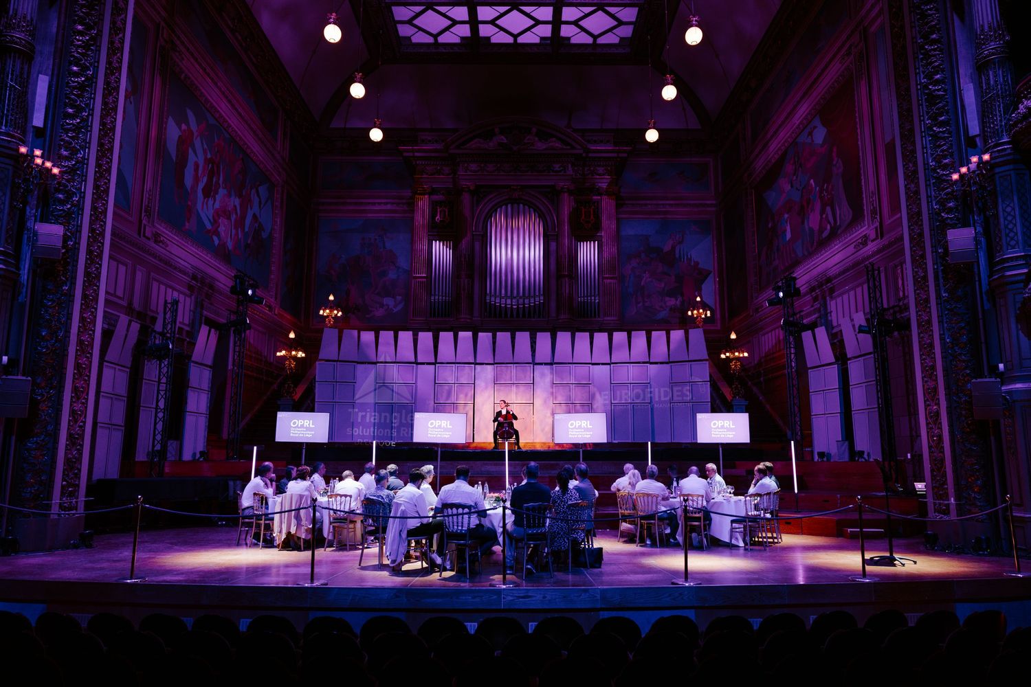 Grand concert hall with purple stage lighting, ornate painted walls, pipe organ, and seated orchestra performers.