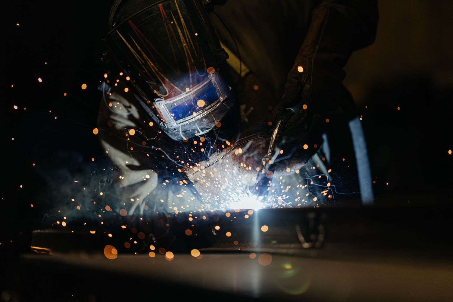 Welder in protective mask creating bright sparks while welding metal in a dark industrial setting.