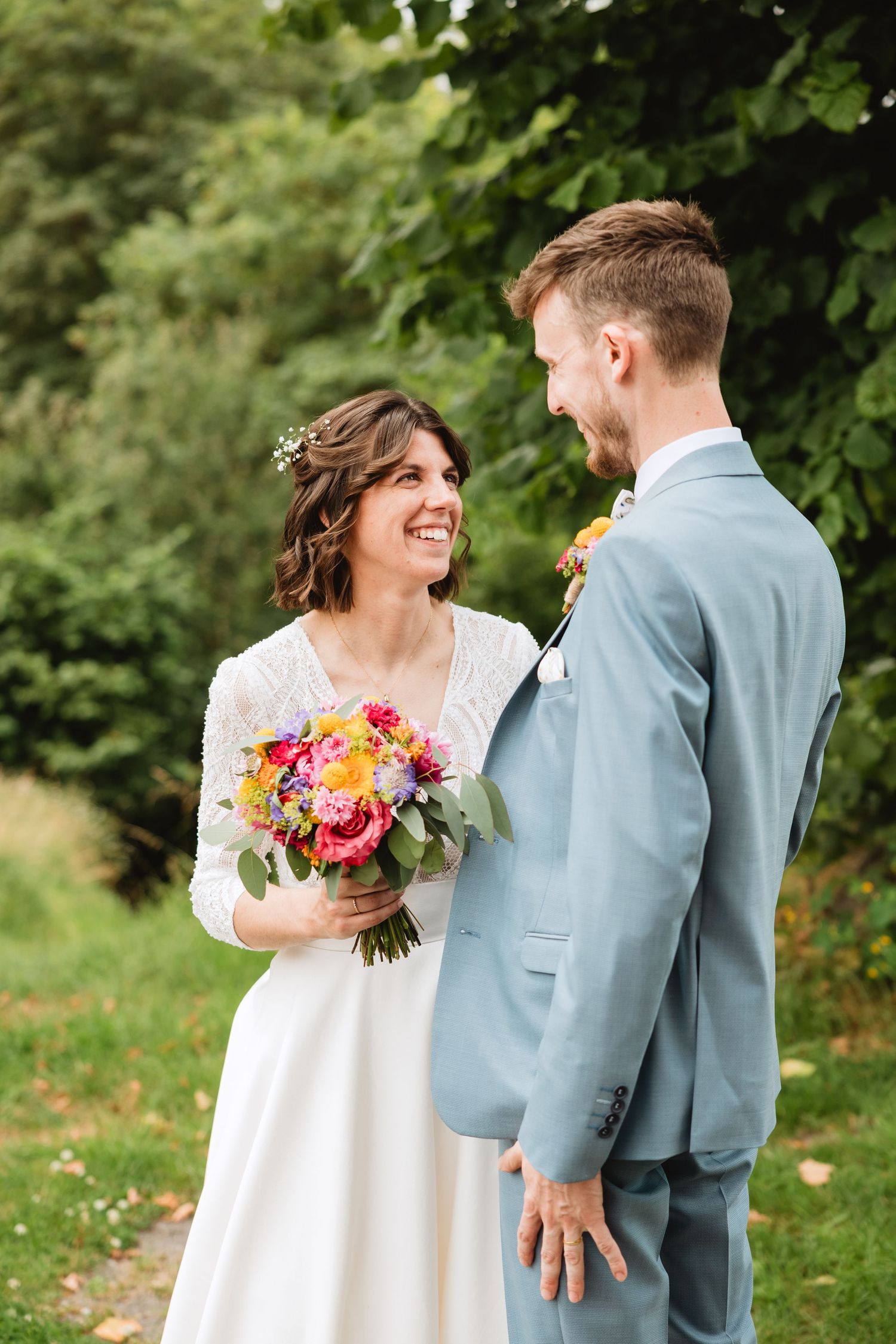 Happy bride holding colorful bouquet smiling at groom in light blue suit in a lush garden setting.