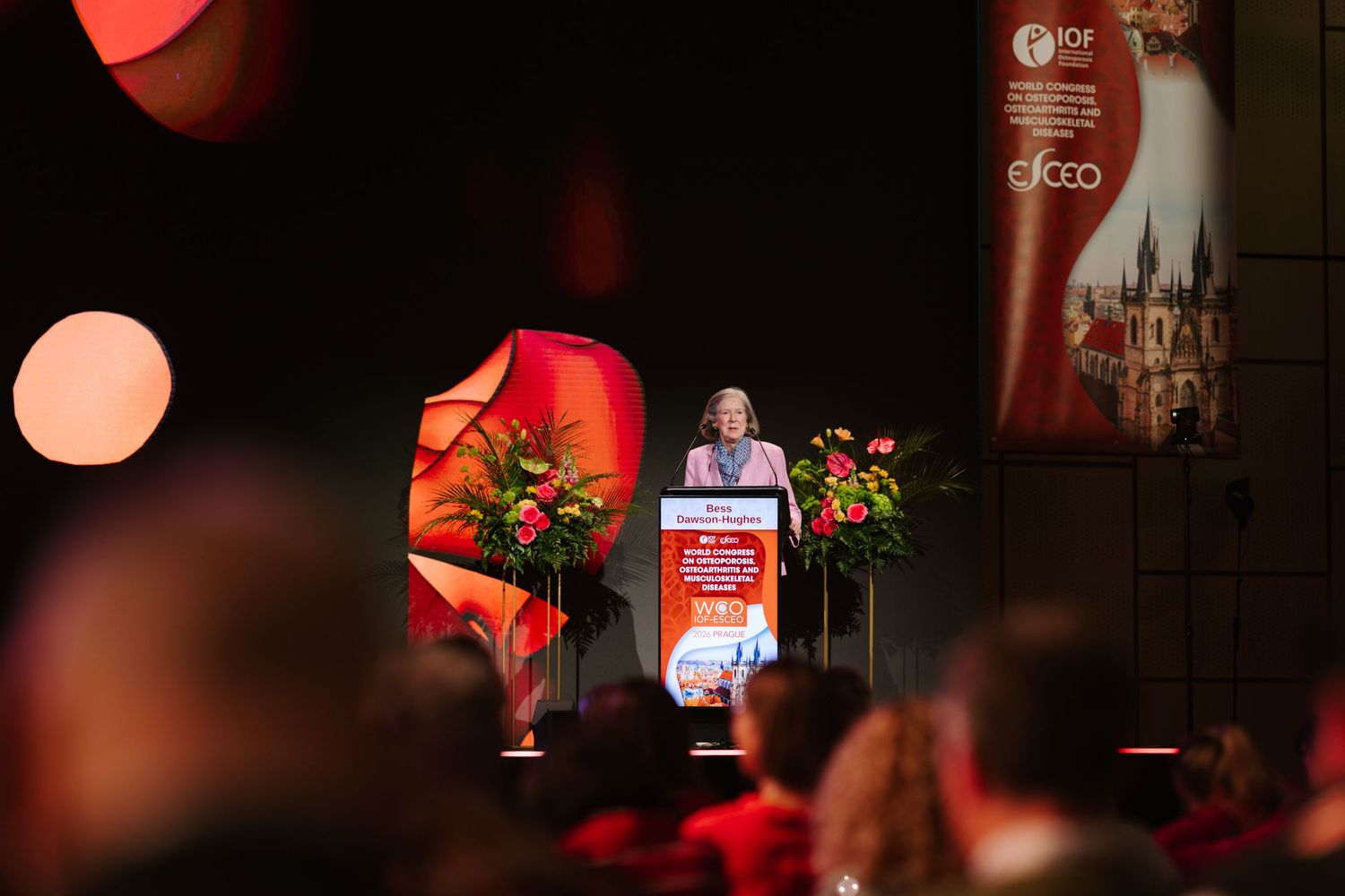 A woman speaks at a podium during an IDF conference, surrounded by red floral arrangements in a dimly lit venue.