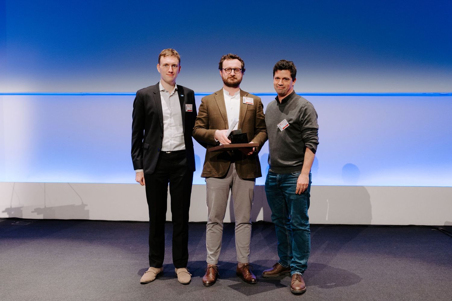 Three professionals pose together on stage at a corporate awards ceremony, with one holding a trophy.