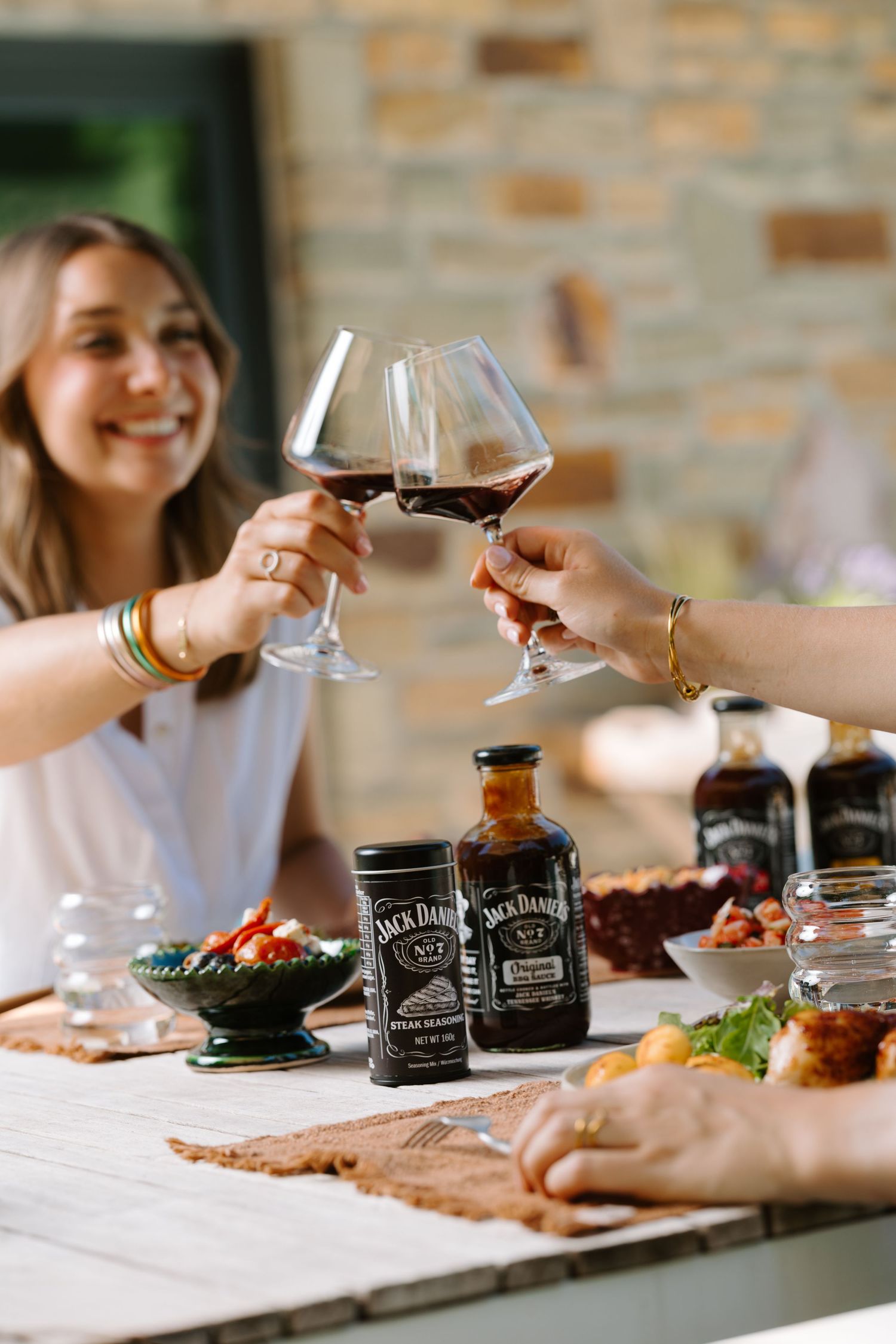 Two women toasting with red wine glasses over a table with Jack Daniel's bottles and food.